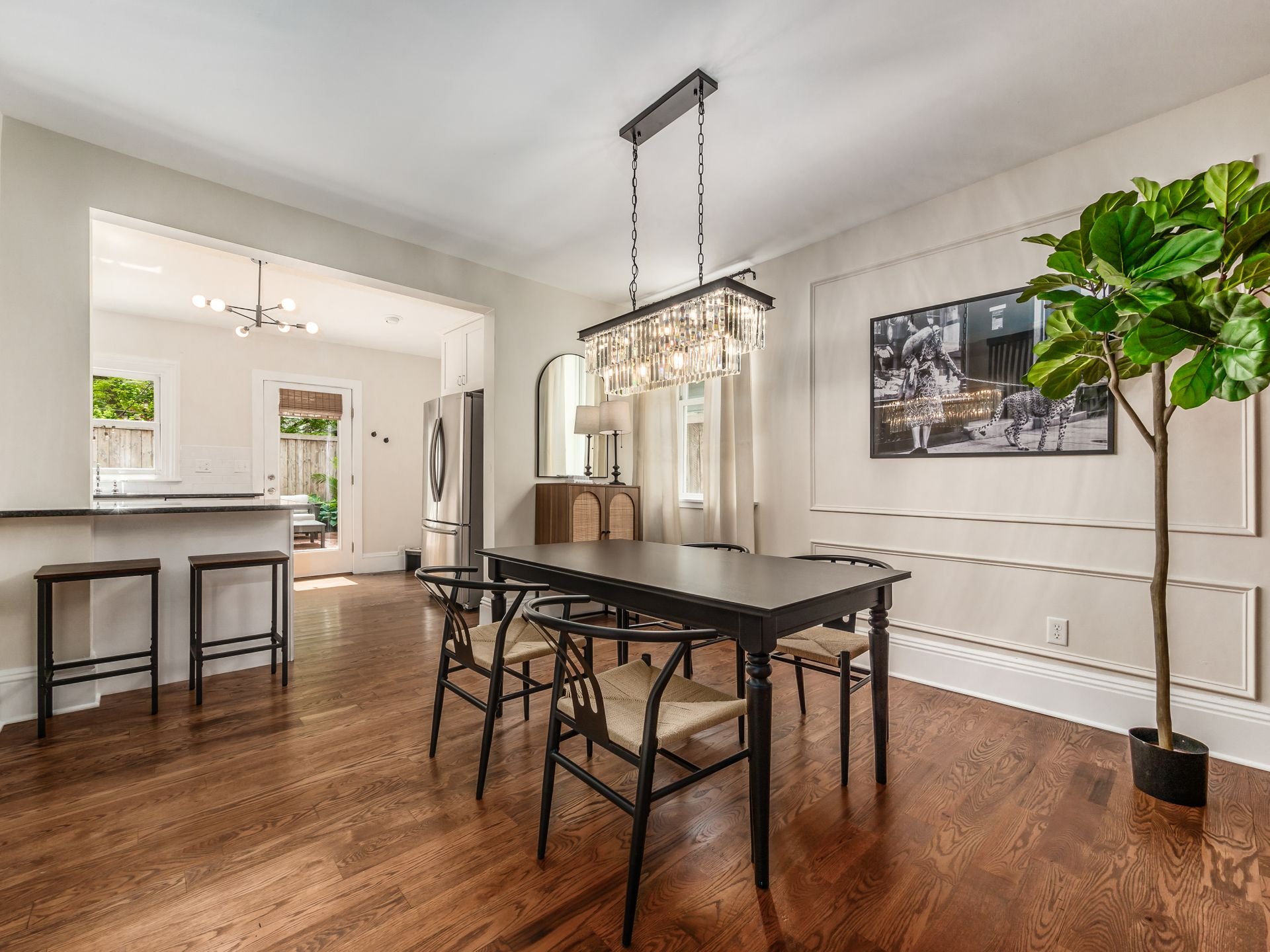 Dining room with wooden floor, black dining table, six black chairs with beige cushions, large chandelier, framed black and white photo of a tiger, tall potted plant, and a cabinet near the window.