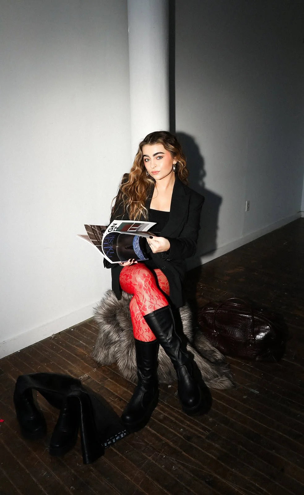 Young woman with wavy hair sitting on a furry stool, wearing a black blazer, red lace fishnet tights, and black knee-high boots, reading a magazine in a minimalistic white-walled room with dark wooden floor.