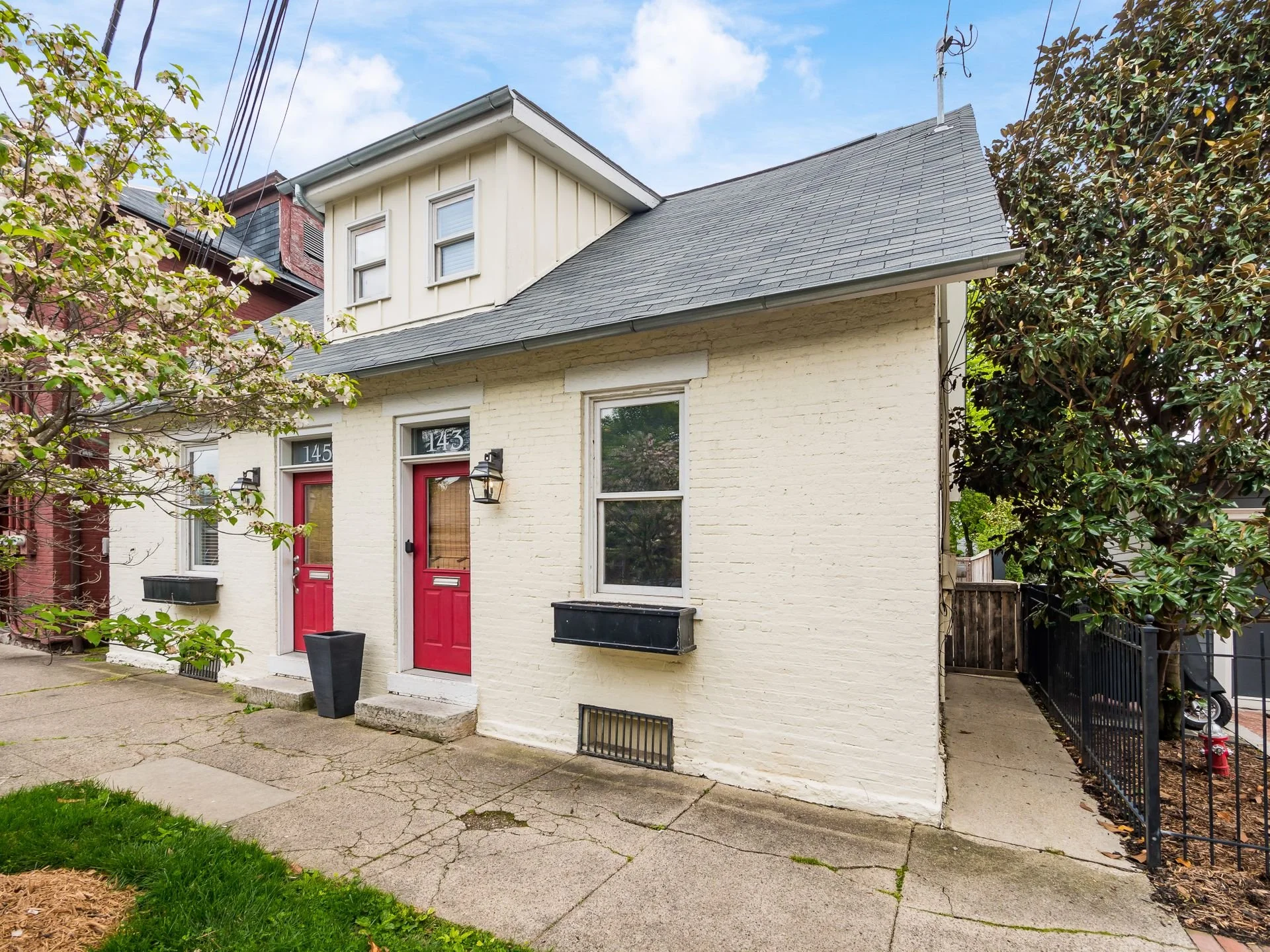 A duplex house with two red front doors, white brick exterior, small porch steps, and black window planters, situated on a cracked concrete sidewalk.