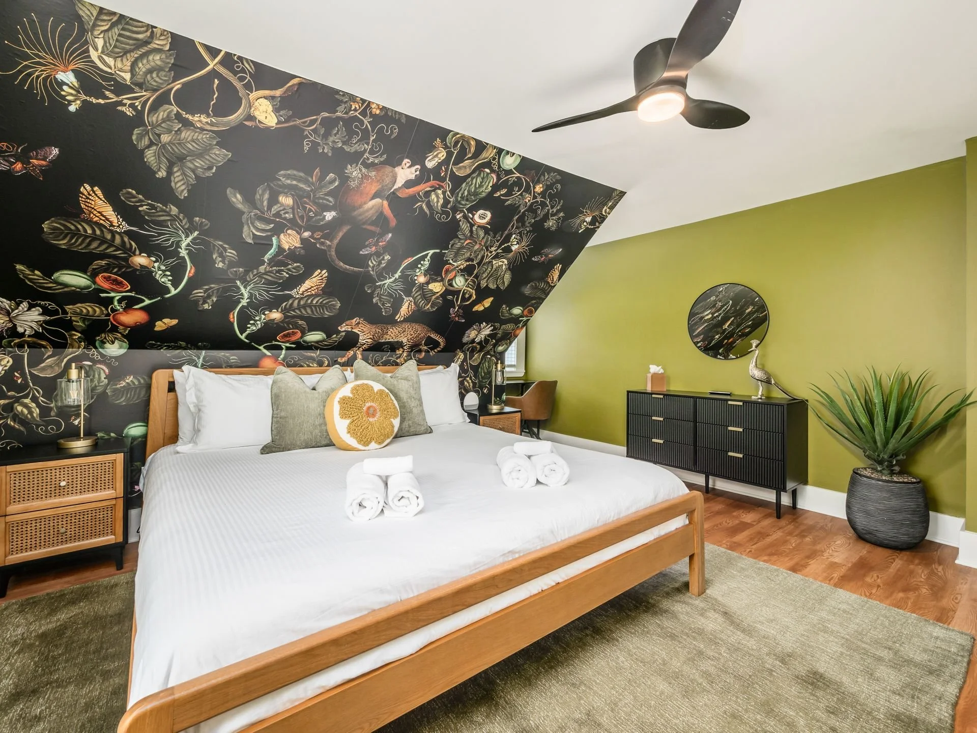 A bedroom with a wooden bed, white bedding, and rolled towels on top. The wall behind the bed has a dark, floral wallpaper mural with animals. To the right, there's a green accent wall with a black dresser, mirror, and decorative items. A large potted plant is in the corner, and a ceiling fan is overhead.