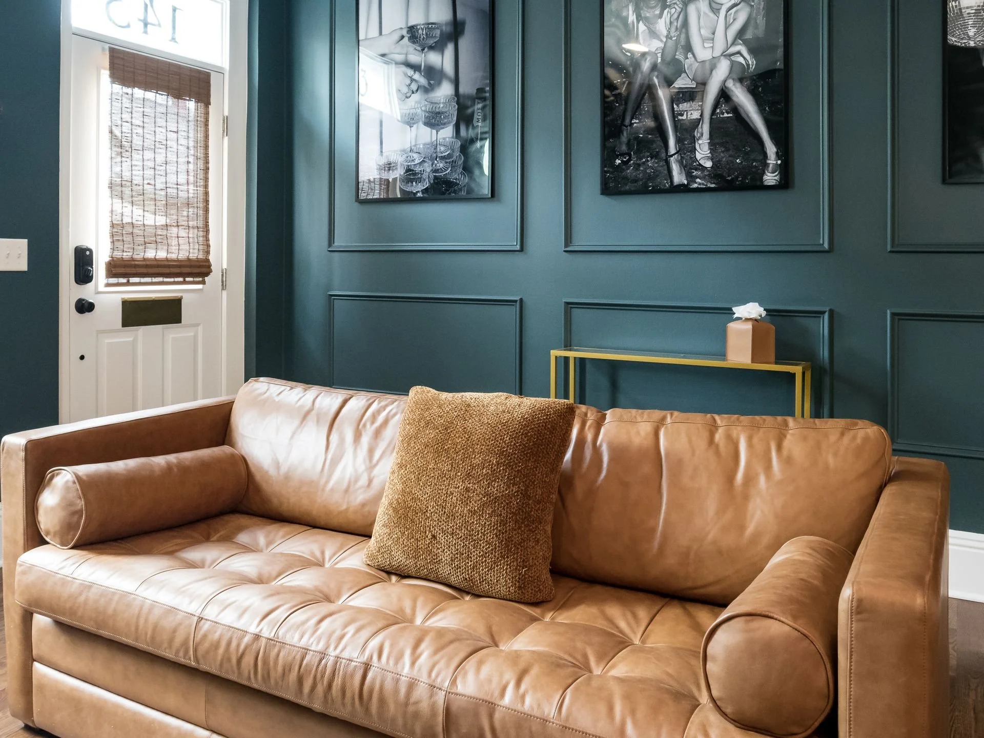 Living room with blue paneled walls, a tan leather sofa with a brown textured pillow, a small yellow side table with tissue box, wall art including a photo of champagne glasses, and a door with a bamboo shade.