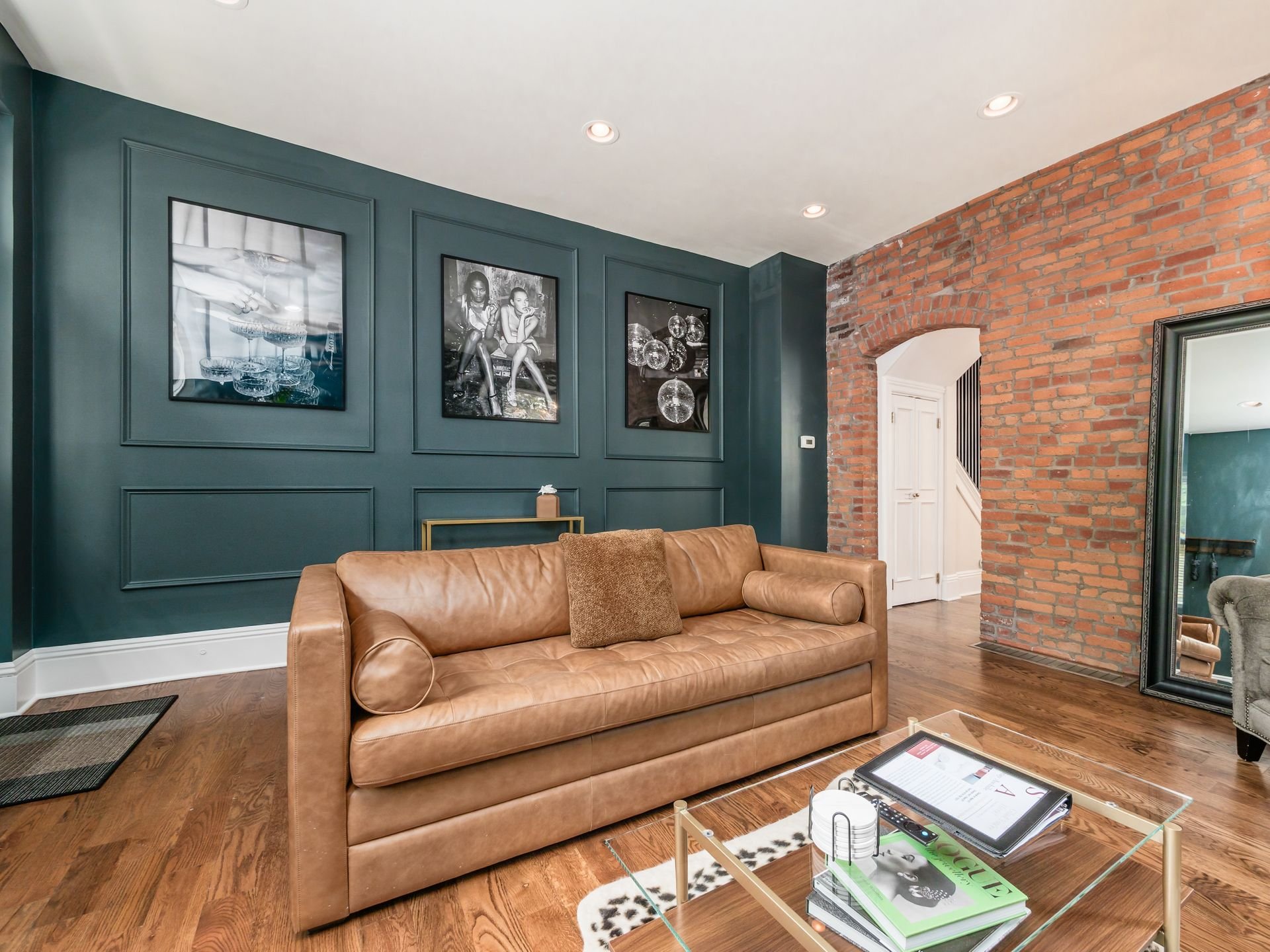 Living room with a tan leather sofa, teal wall with three framed black and white photos, exposed brick wall, hardwood floor, large mirror, glass coffee table with magazines and a tablet, and recessed ceiling lights.