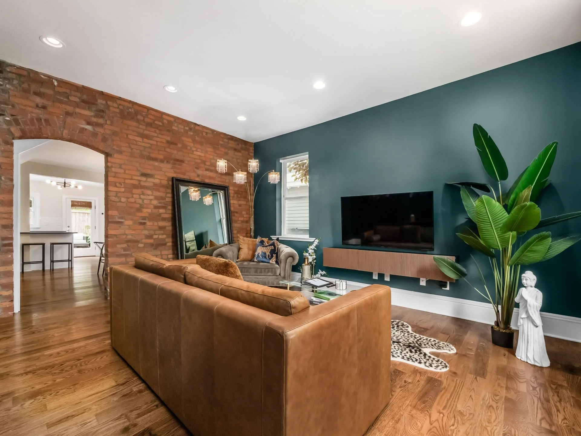 Living room with a brick wall, teal accent wall, brown leather sofa, large mirror, TV, large green plant, decorative statue, window, hardwood floors, and modern ceiling lights.