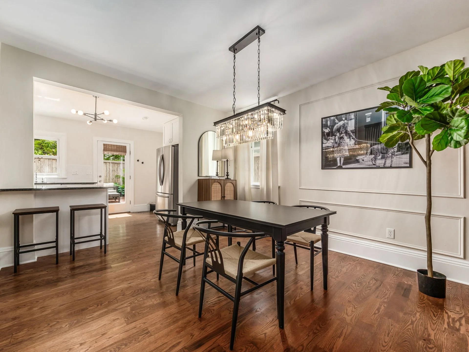 Dining room with black table, four chairs, and chandelier, adjacent to kitchen with two bar stools and refrigerator, large potted plant, black and white photograph on wall, hardwood floors, and window with curtains.