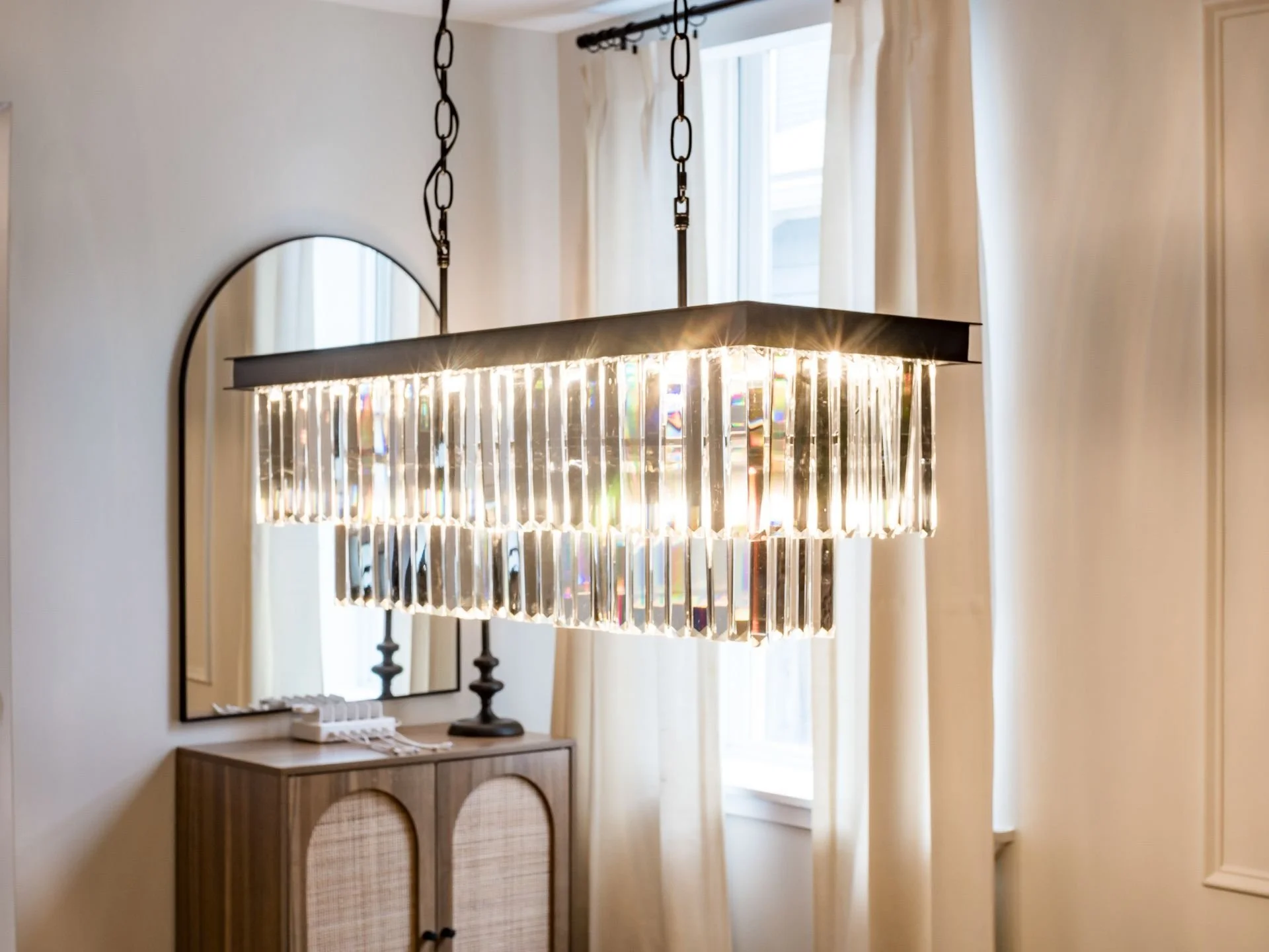 Modern rectangular chandelier with hanging crystals, glowing brightly in a room with white walls, a window with curtains, a mirror, and a wooden cabinet.