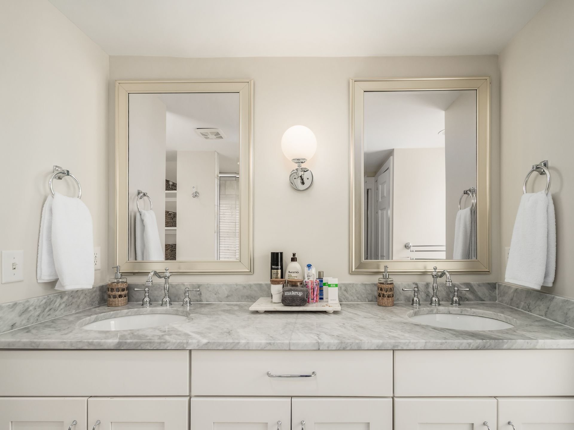 Double bathroom vanity with marble countertops, two sinks, silver faucets, two mirrors, a wall-mounted light fixture, and personal care items on a tray in the center.