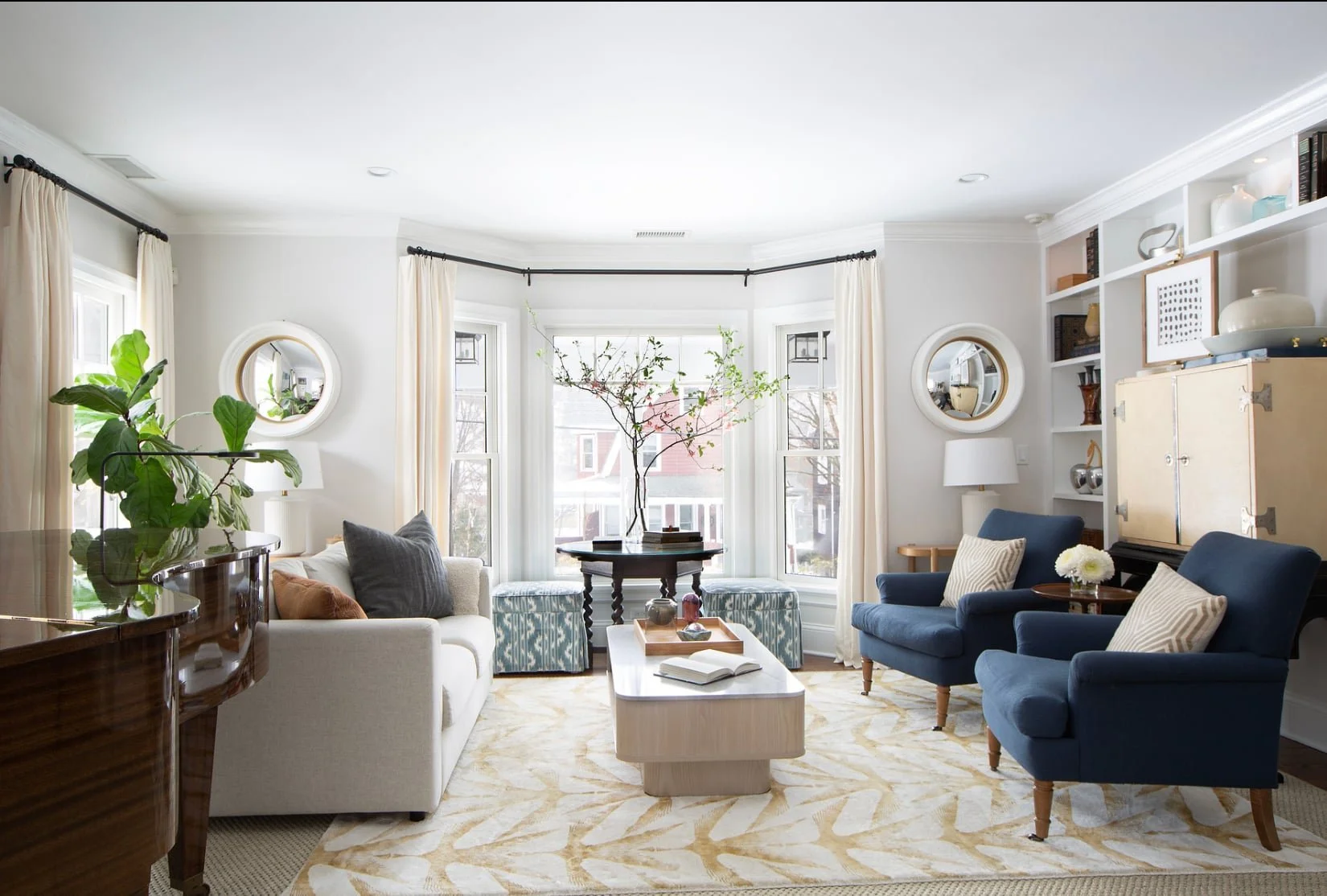 Bright living room with large bay windows, beige curtains, cream sofa with pillows, two blue armchairs with pillows, a coffee table with a book and decorative items, a small round table with a plant, and built-in white shelves with decor items.