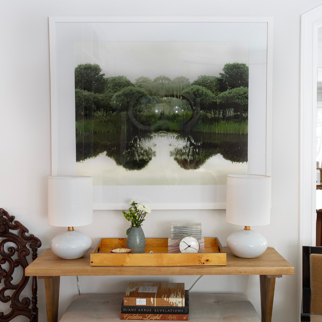 A living room wall with a large framed photograph of a green forest and its reflection in water, flanked by two white table lamps on a wooden console table with decorative items and books.