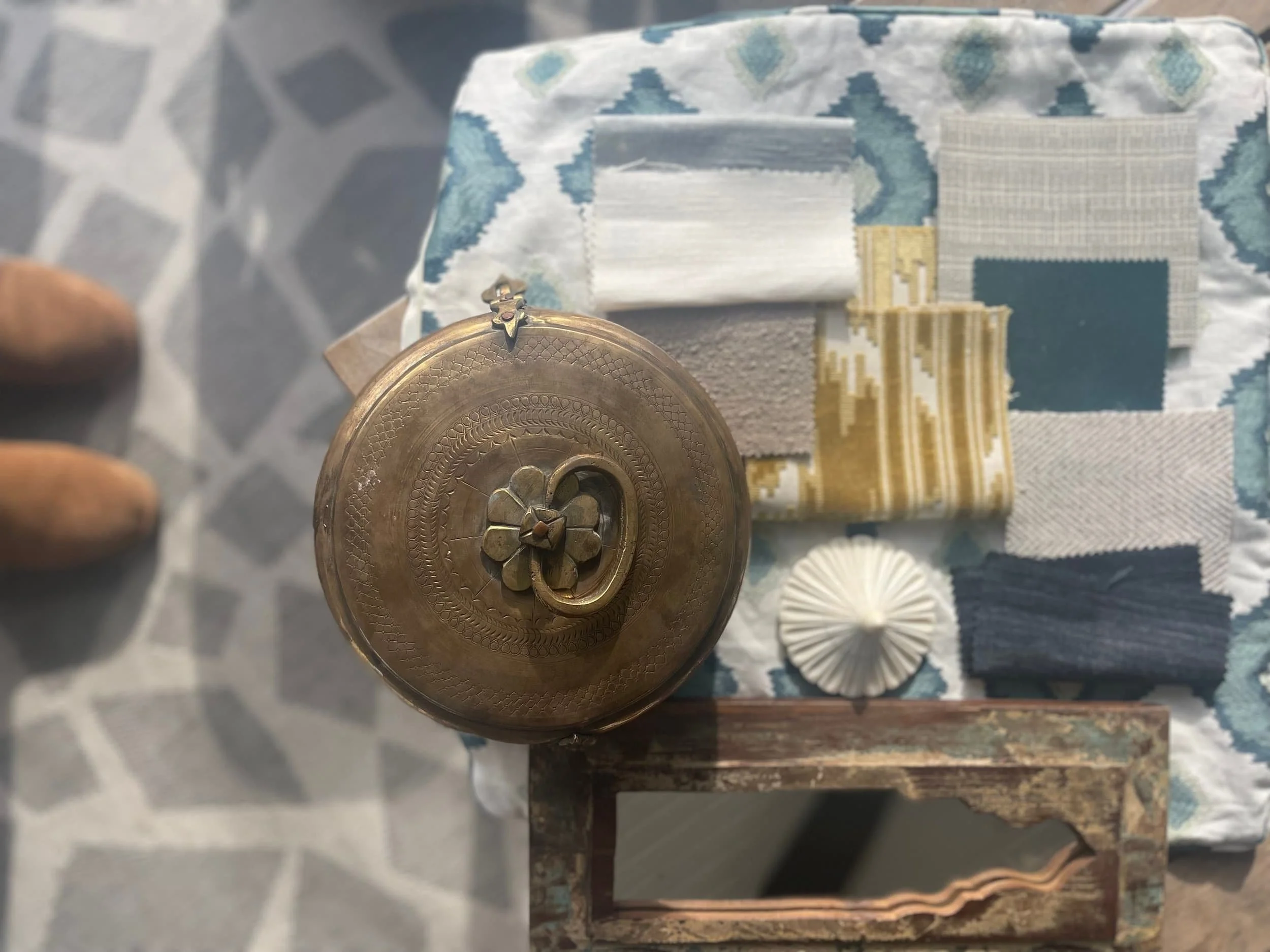 Various fabric swatches laid out on a table, with a brass decorative object with a four-leaf clover and heart design, a small round white decorative item, and a rustic mirror underneath.