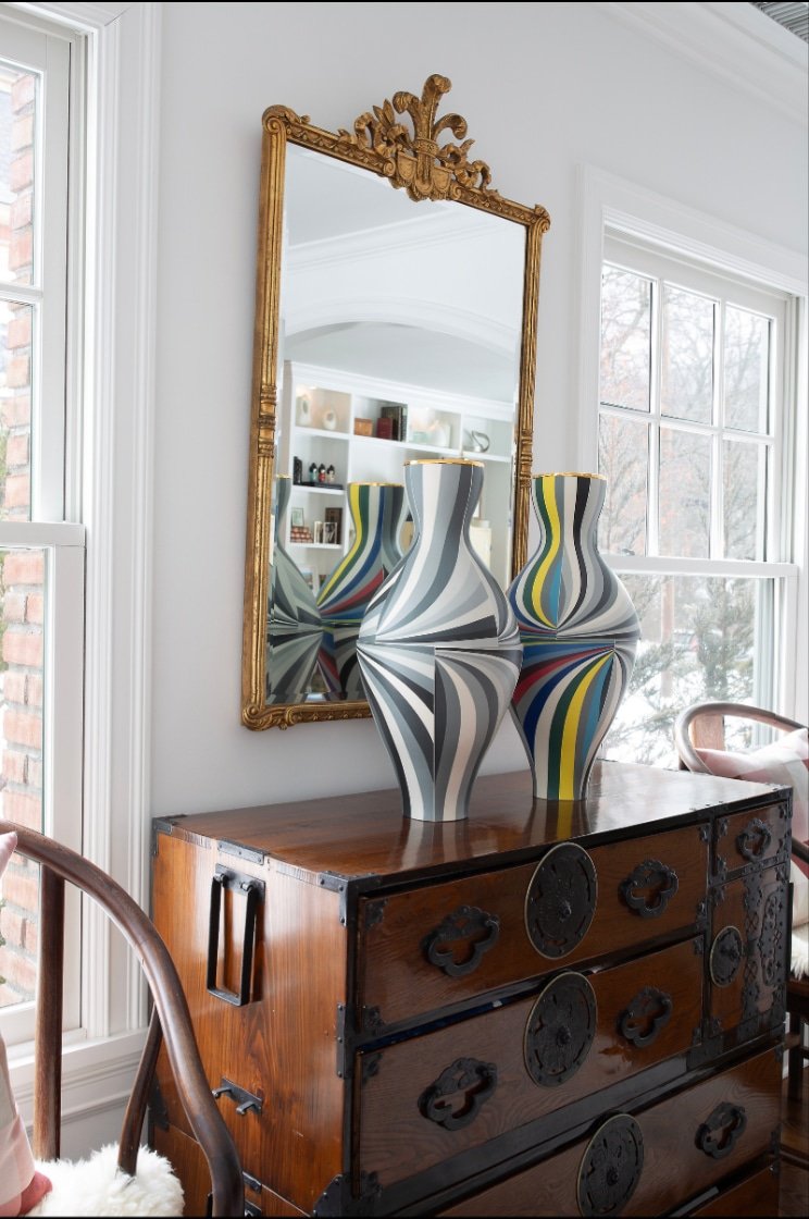Two abstract striped vases with black, white, gray, yellow, green, and red patterns are placed on a wooden dresser beneath a large ornate gold-framed mirror. The dresser is against a white wall with large windows to the right, revealing a room with b