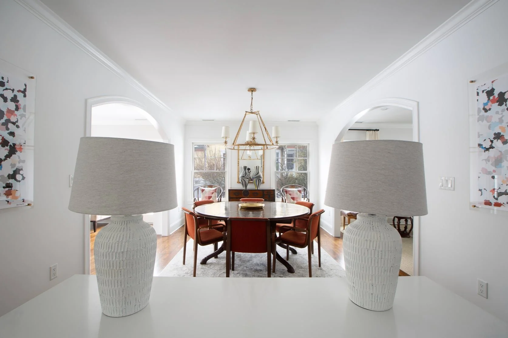 A dining room with a round wooden table surrounded by six red upholstered chairs, two colorful vases and a chandelier hanging above, viewed from a space with two white lamps on a counter in the foreground.