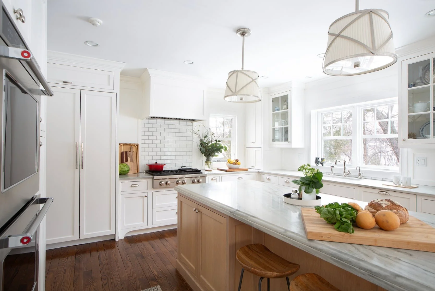 Bright white kitchen with wooden flooring, large island with a marble countertop, and window with natural light. Kitchen features white cabinets, a white subway tile backsplash, and pendant lighting. Items on island include potted plant, bread, and v