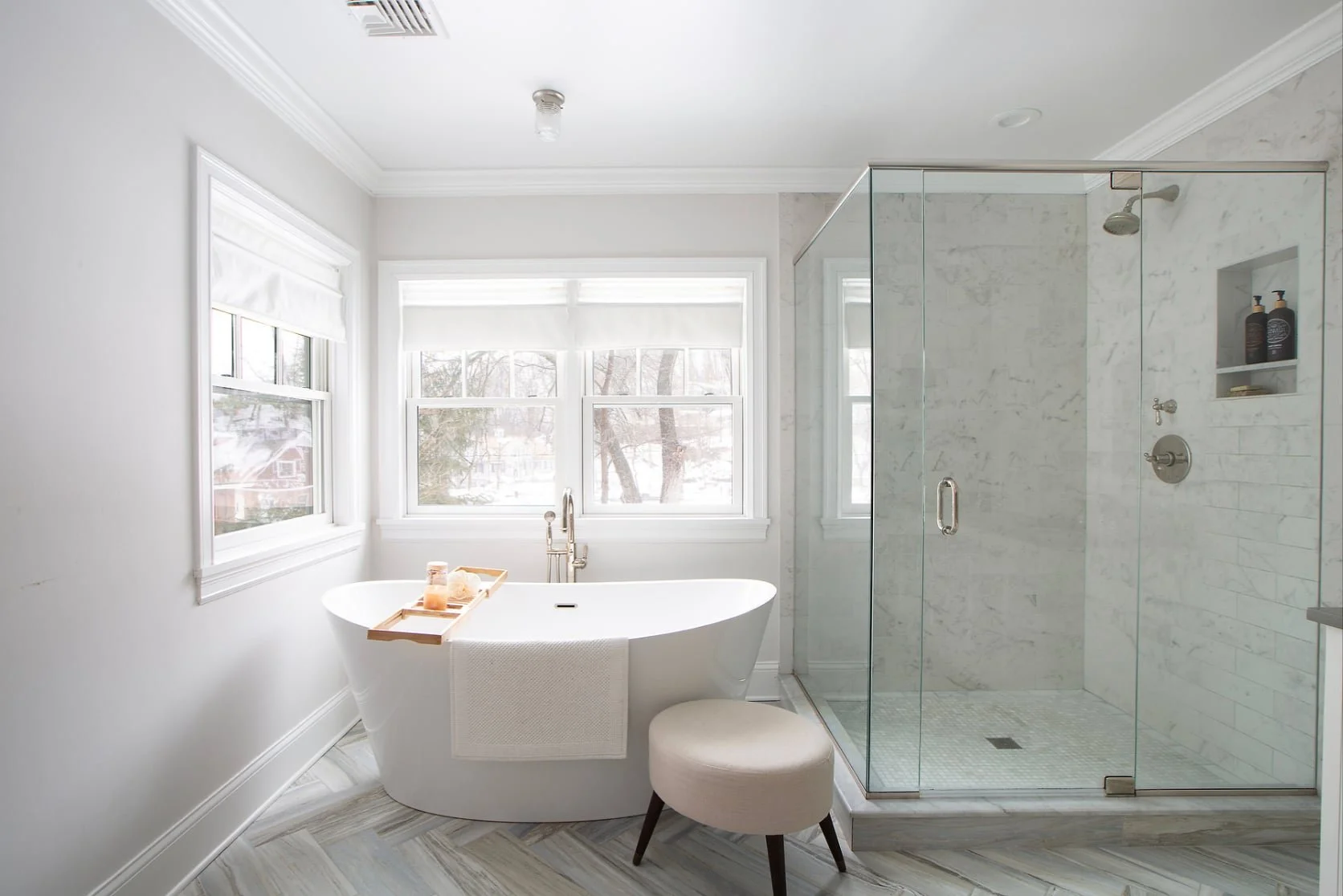 Bright bathroom with a white freestanding bathtub on the left under three large windows with white blinds, and a glass-enclosed shower on the right with marble tiles, a showerhead, and built-in shelves.