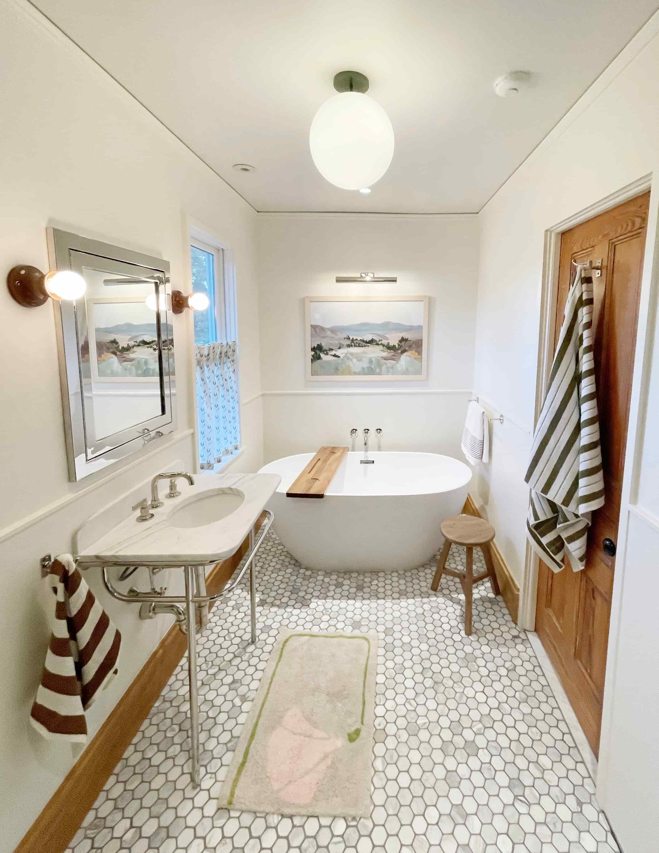 A cozy bathroom featuring a bathtub with a wooden tray, a vintage-style sink, striped towels, a small stool, hexagon tile flooring, and framed landscape art on the walls.