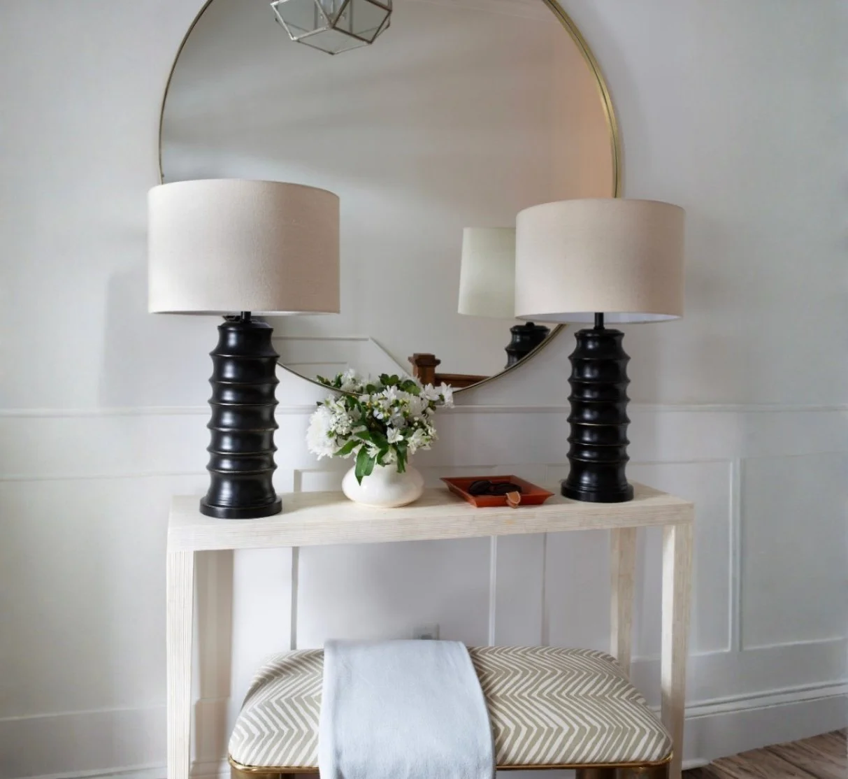 A white console table with two black lamps with beige lampshades, a small white vase with white flowers, and a small tray with jewelry, positioned in front of a large round mirror on a white wall.