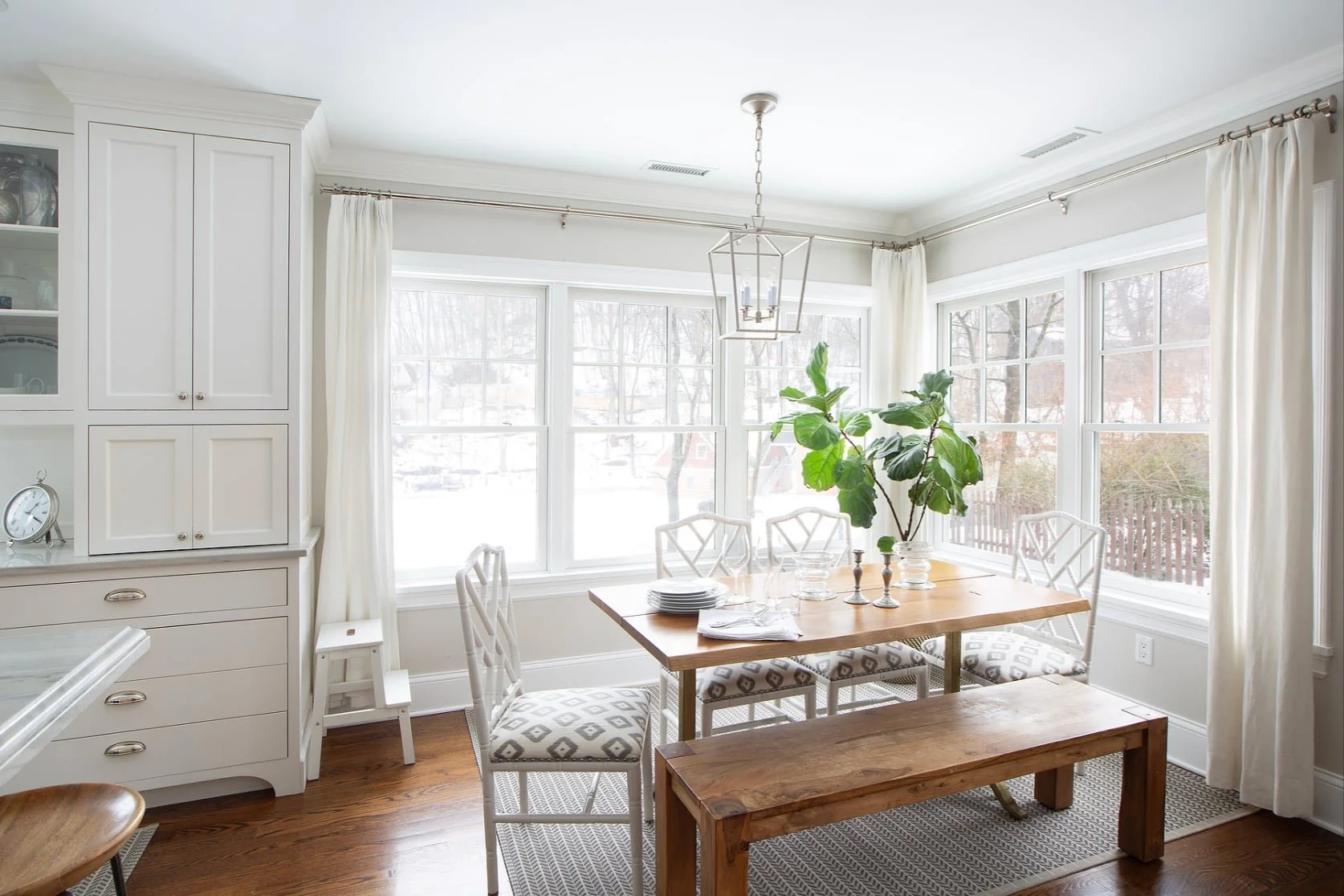 Bright dining room with large windows, white walls, a wooden table, white chairs with patterned cushions, a wooden bench, a large green plant, and a modern light fixture.