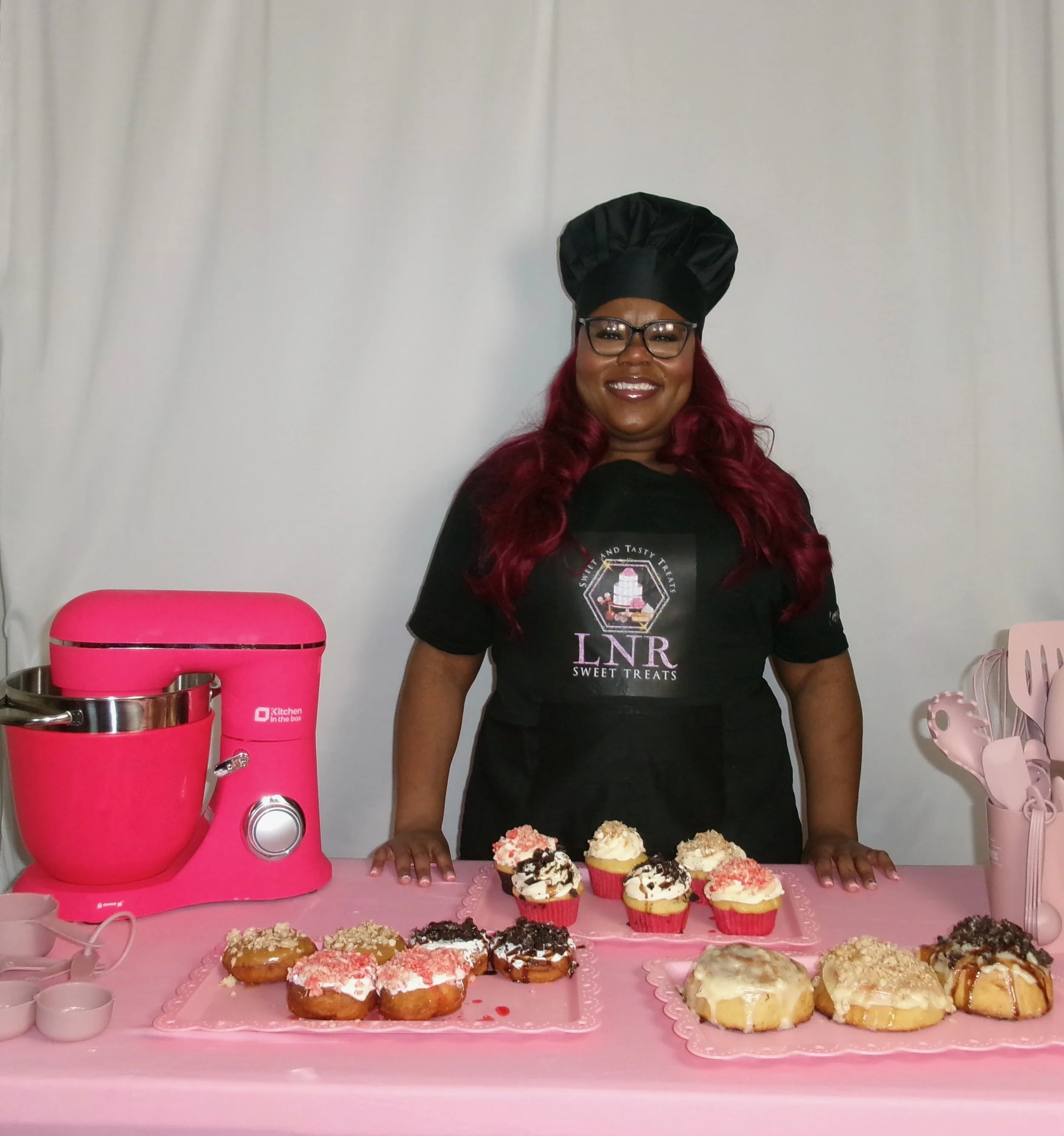 Woman in a chef's hat and apron standing behind a table with assorted decorated cupcakes and donuts, a pink mixer, and pink utensils.