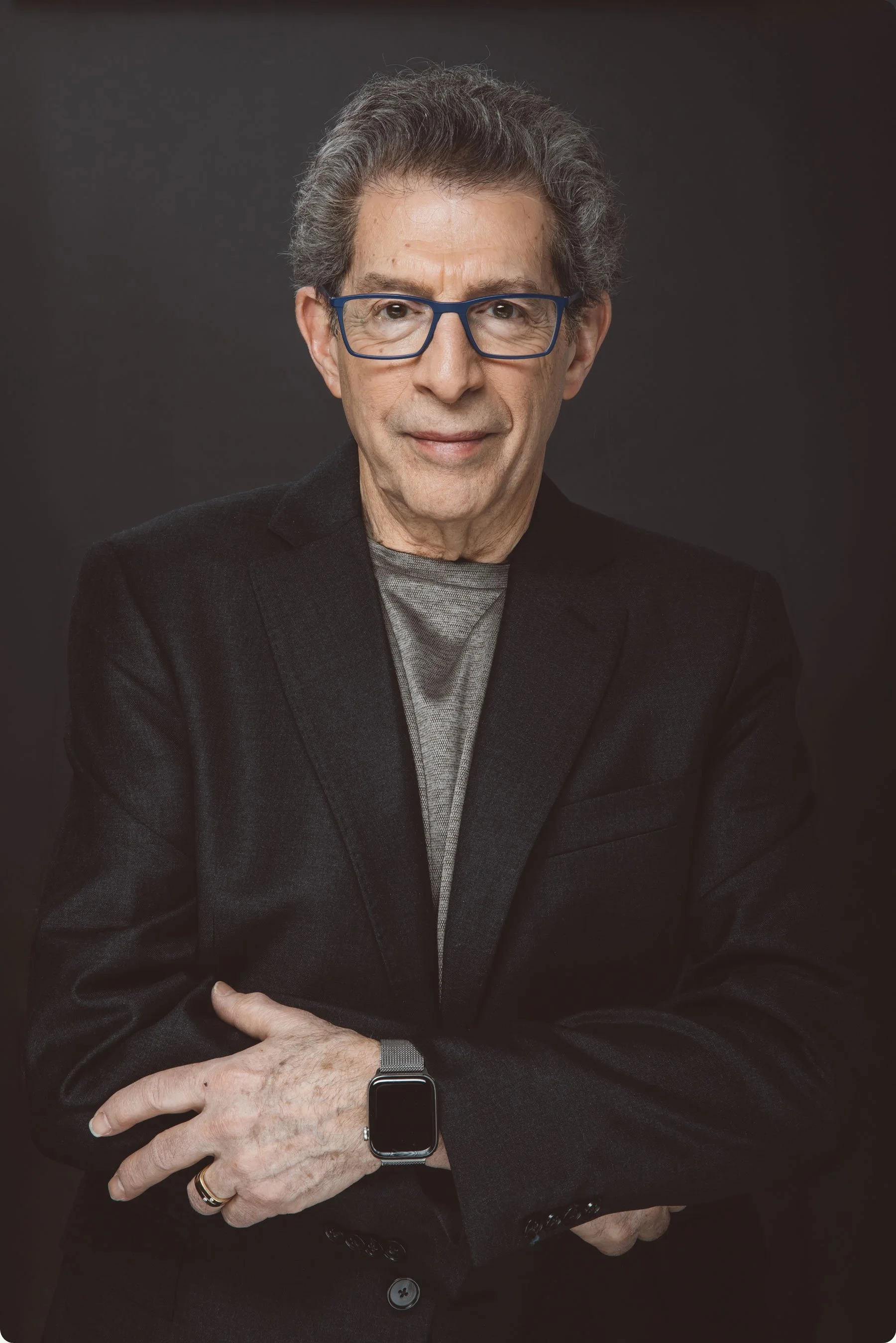 A portrait of Mark Shekter wearing a black blazer and gray shirt, standing against a dark background.