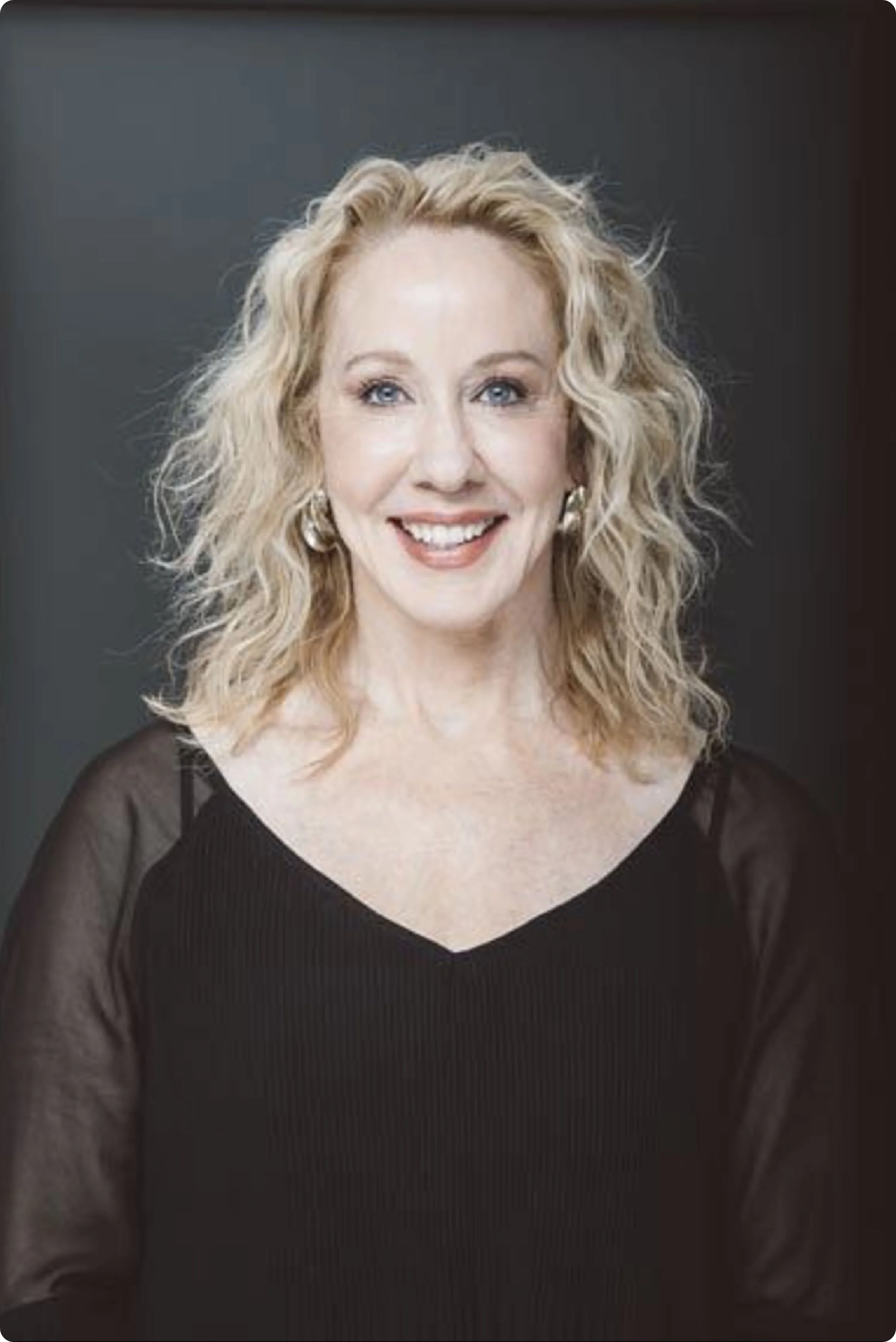 A portrait of Janell who is smiling with wavy blonde hair, wearing a black top with sheer long sleeves, earrings, against a dark gray background.