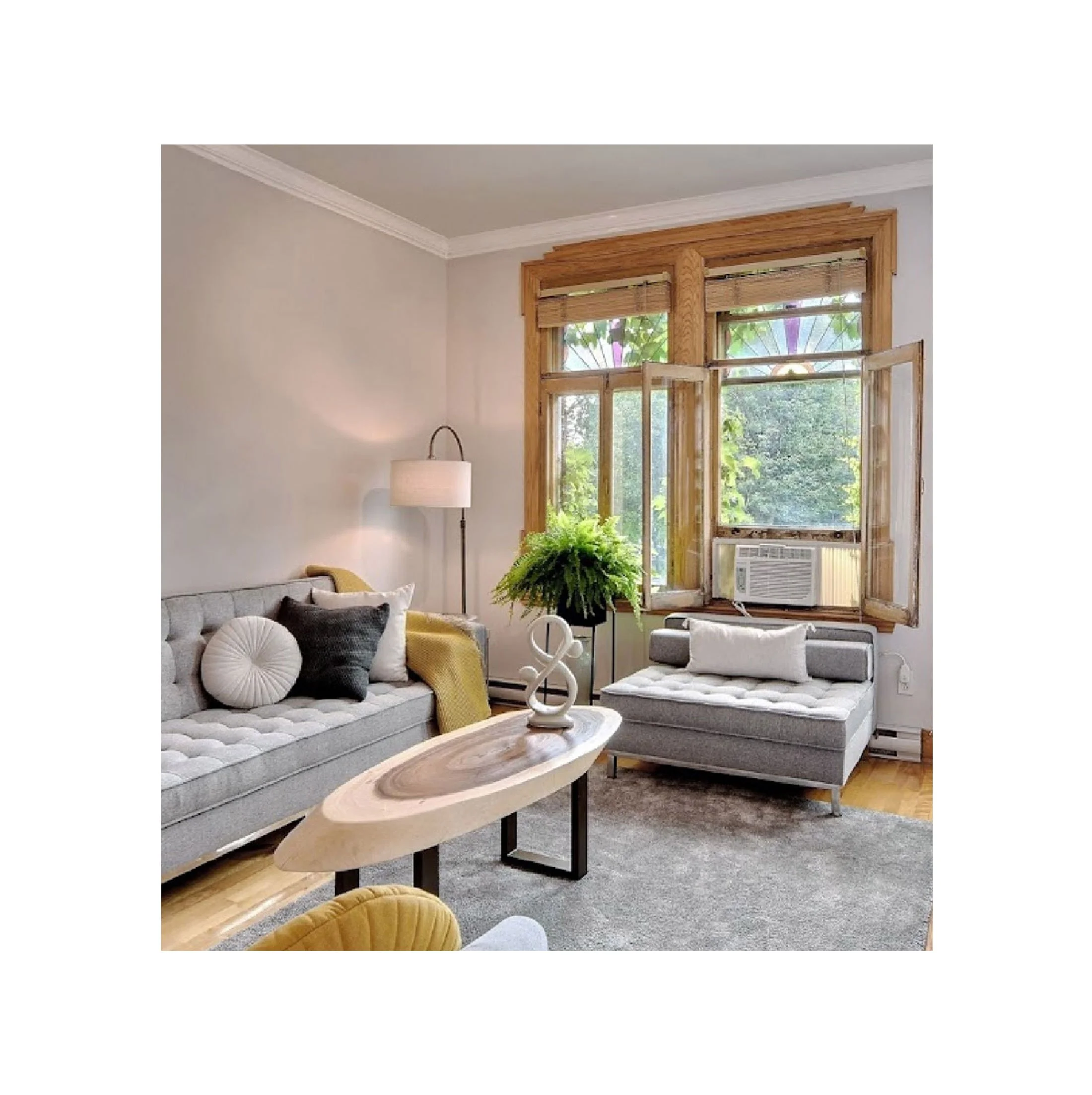 Living room with large wooden-framed windows, gray sofa with pillows, a modern coffee table, and a tall lamp, with lush greenery visible outside.