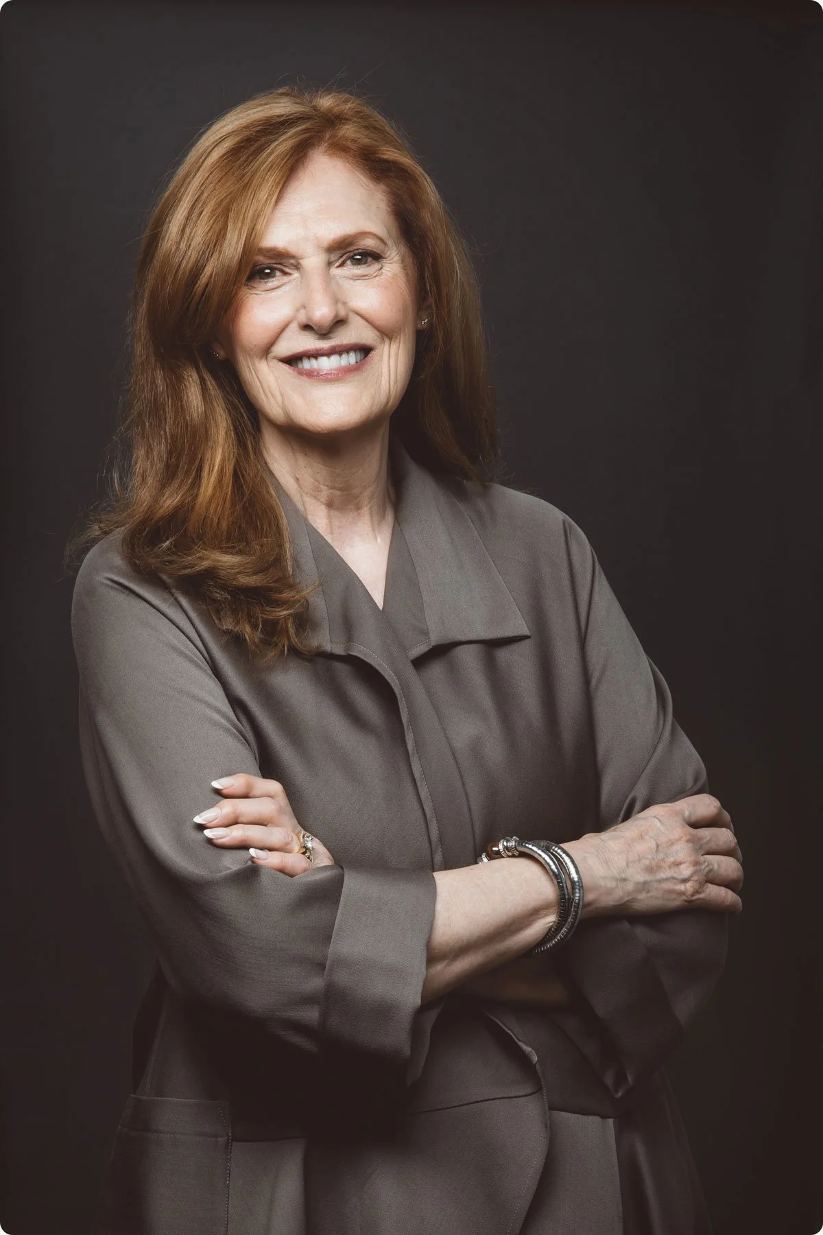 A portrait of Nancy Trites Botkin who has long auburn hair and is smiling, standing with her arms loosely crossed against a dark background.