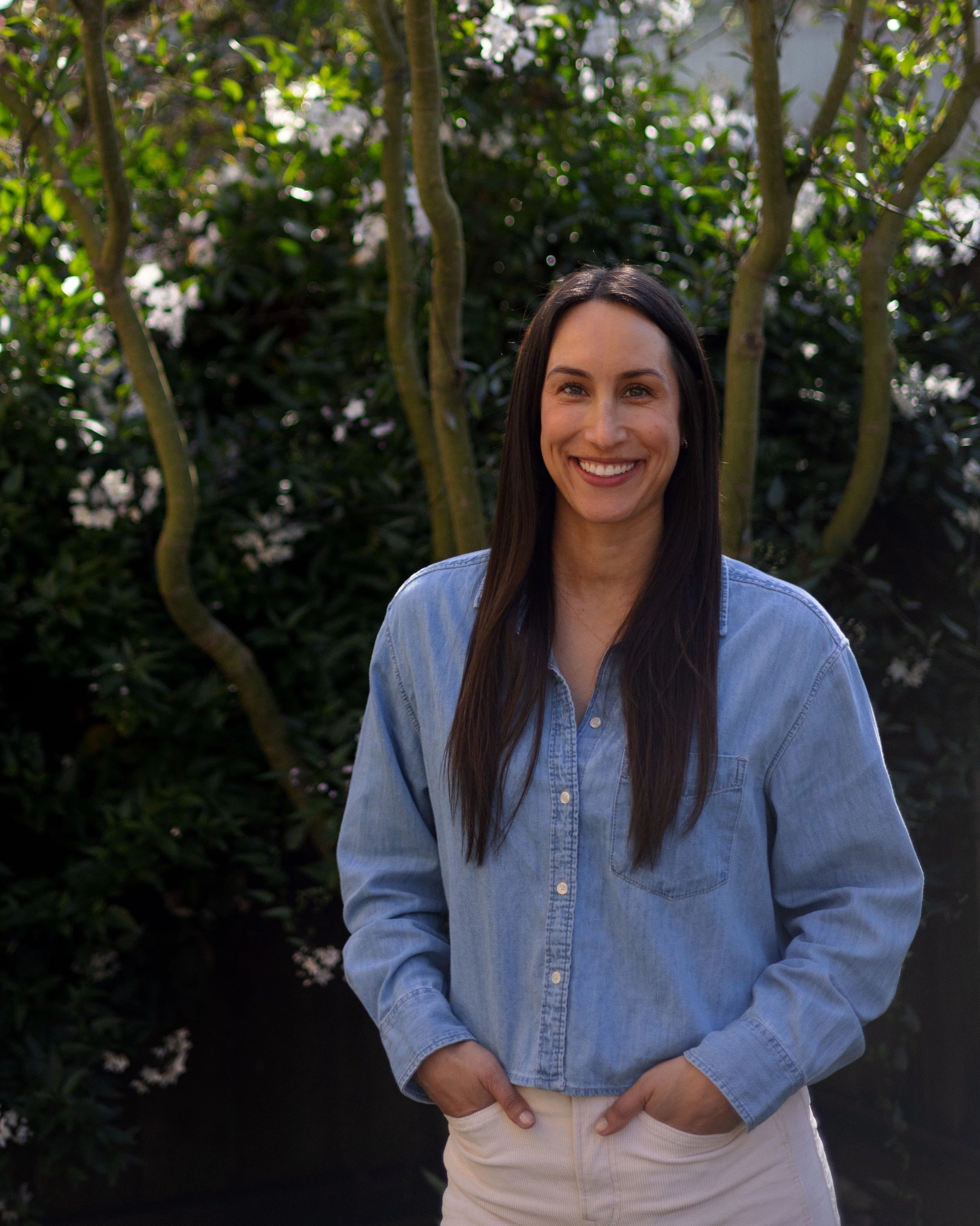 A woman with long dark hair, wearing a light denim shirt and white pants, smiling with her hands in her pockets in front of green trees.