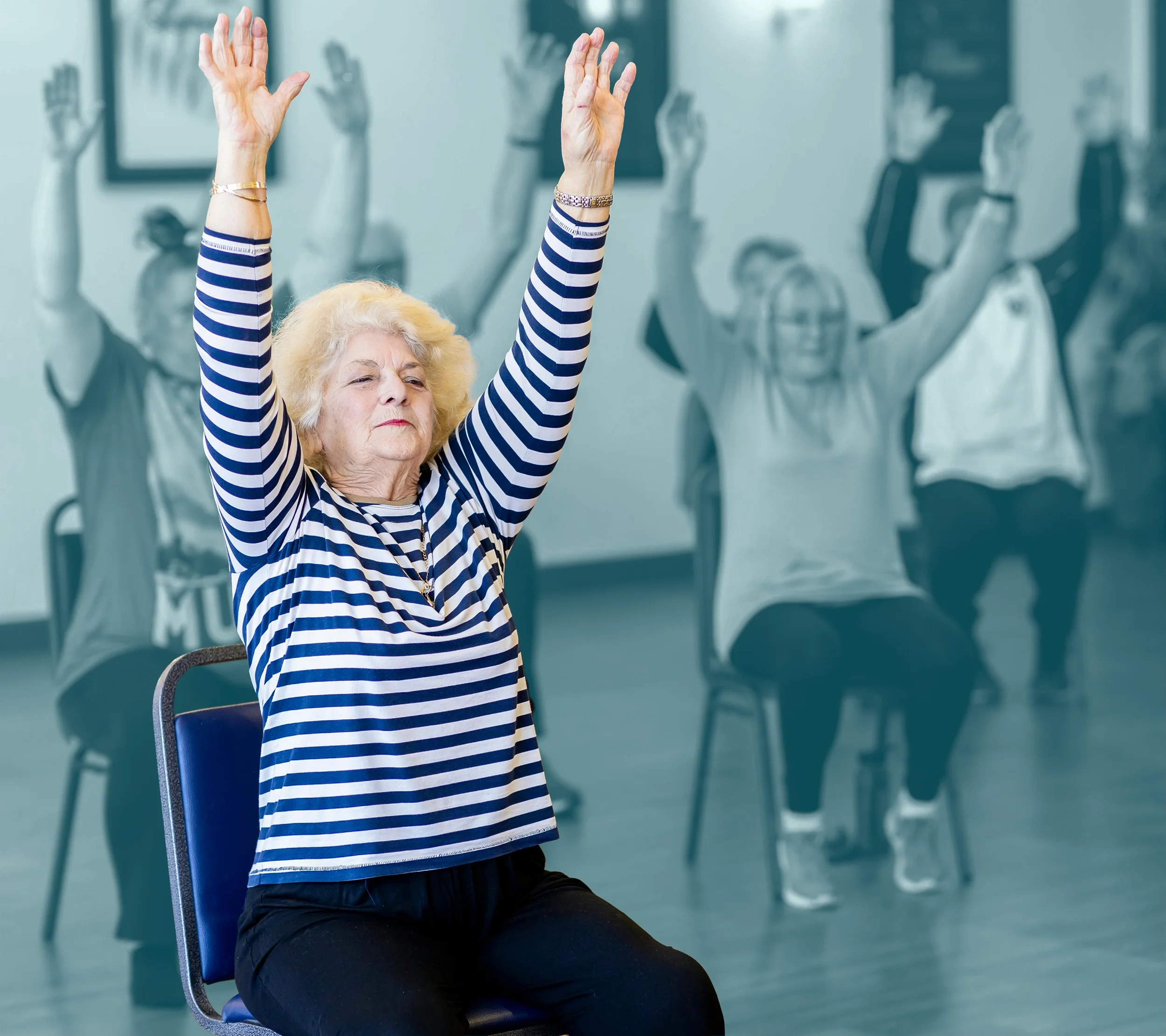 An elderly woman with blonde hair, wearing a striped long-sleeve shirt, is sitting on a chair with her eyes closed and arms raised. Behind her, there are some blurred people also raising their arms, in what appears to be a group exercise or yoga class.