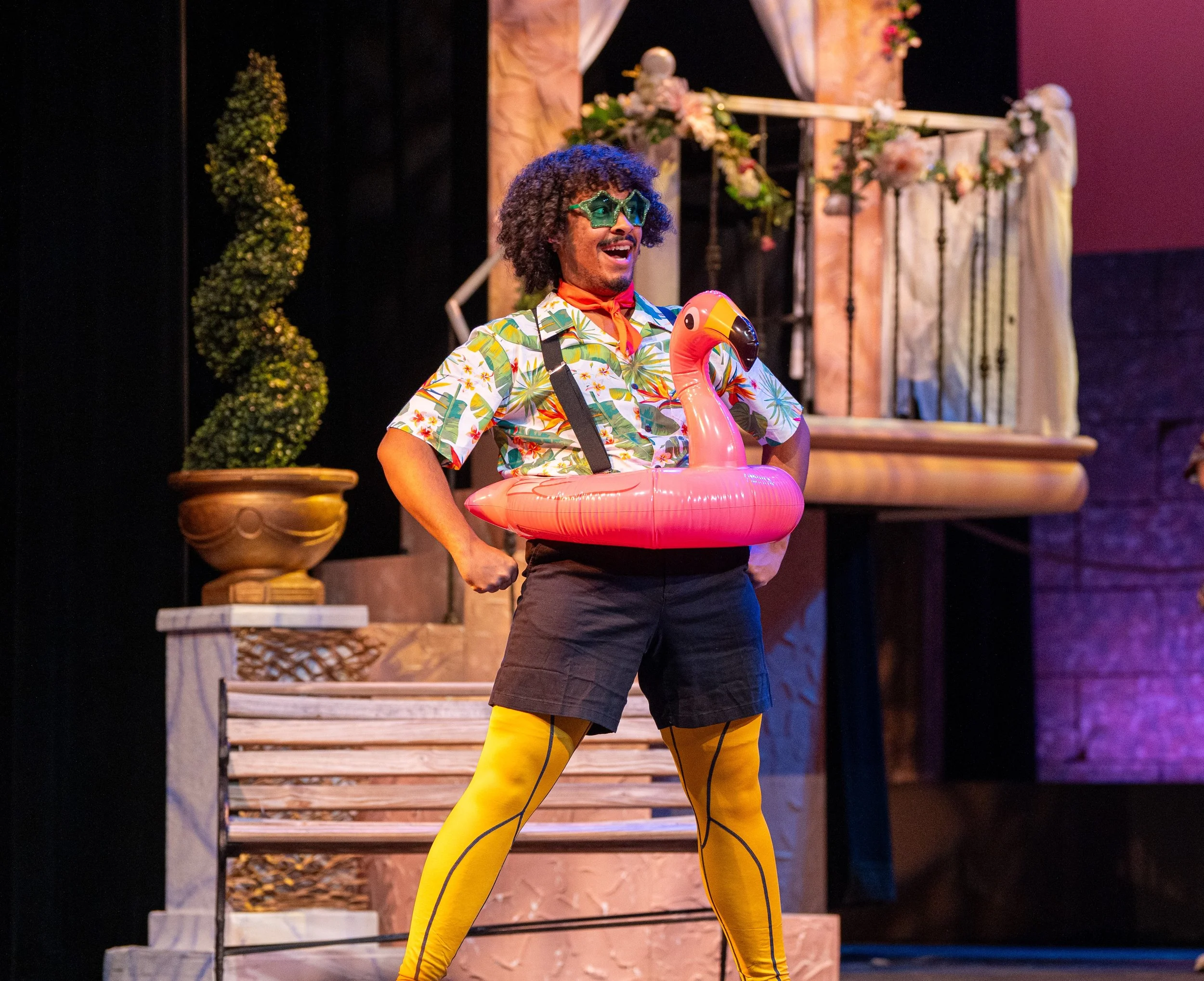 Performer singing 'Count Malvolio' from 'Twelfth Night' musical adaptation wearing bright yellow leggings, a Hawaiian shirt, and sunglasses, with a pink flamingo pool float around his waist, on stage with decorative flowers and greenery.
