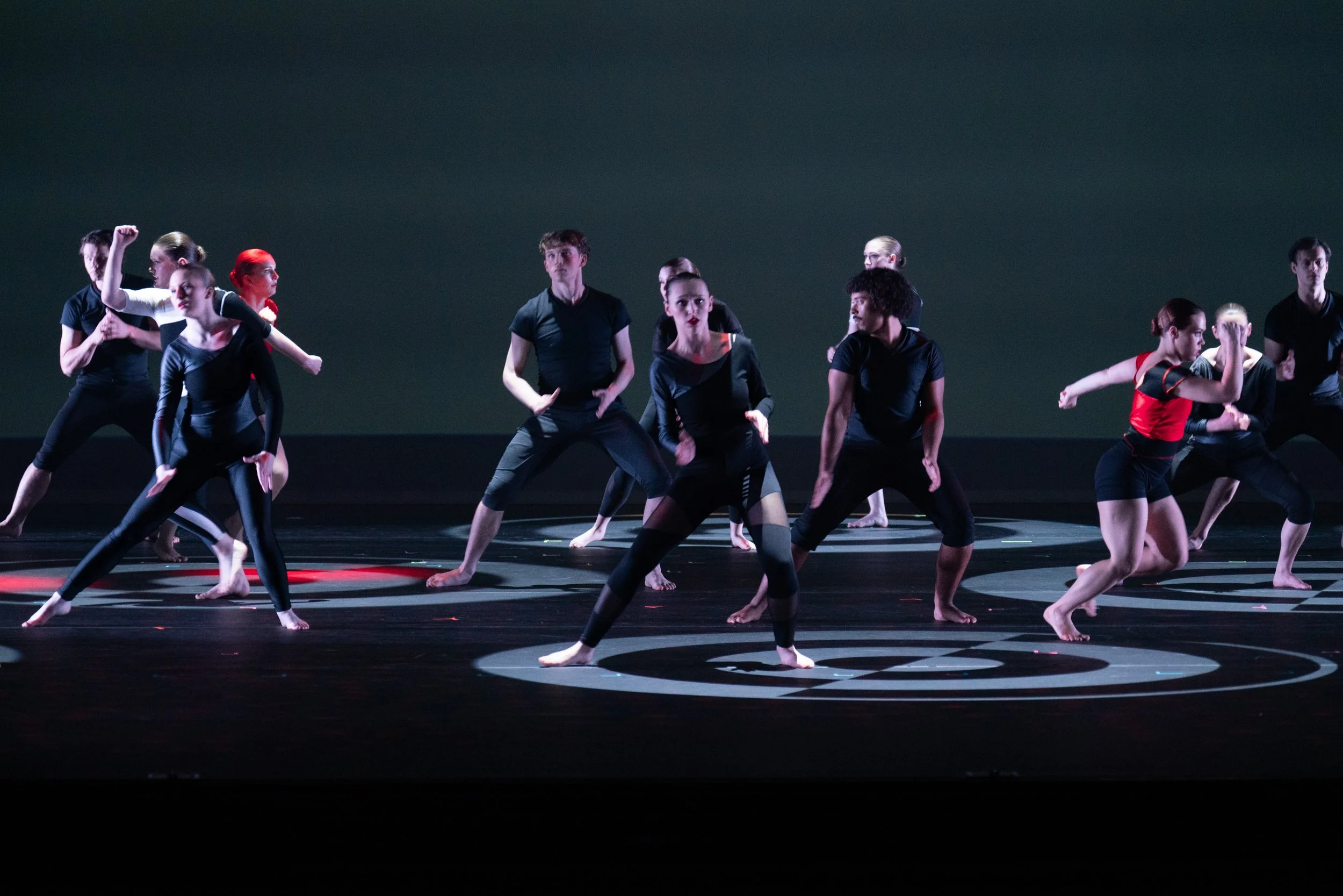 Group of dancers performing on stage in black and red costumes, with various dance poses and expressions.