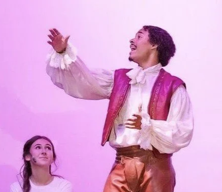 A performer dressed in a colorful costume on stage, mid-performance, with a woman seated in front watching.