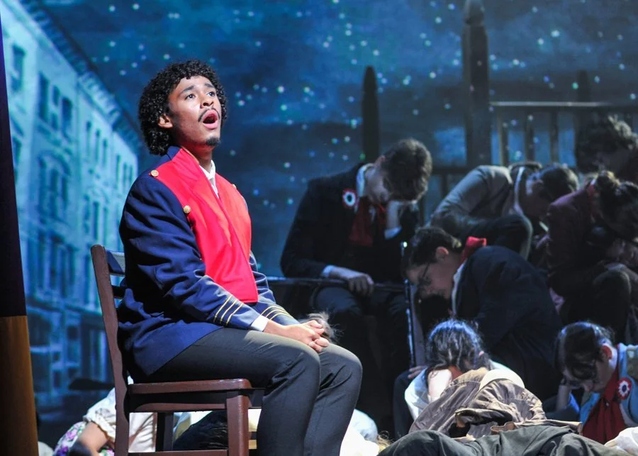 A musical performance of 'Bring Him Home' from 'Les Miserables' with actors dressed in historical costumes on stage, with one actor in a military-style uniform sitting on a chair, singing, while others are sleeping in the background.