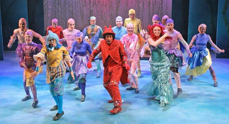 The cast of the musical 'The Little Mermaid' with Sebastian front performing 'Under the Sea' on stage with colorful sea creature costumes.