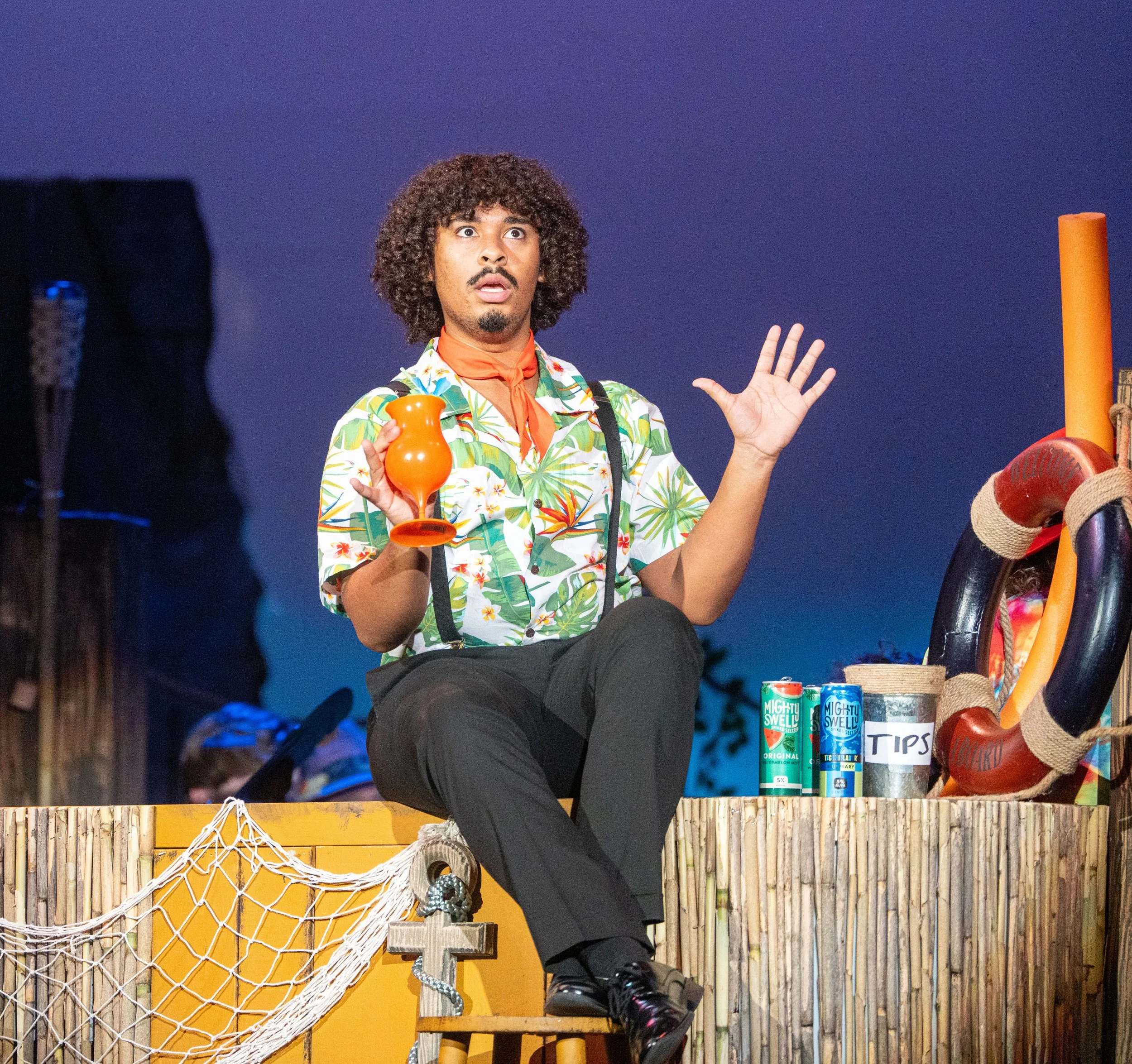 A man with curly hair dressed in a tropical shirt, orange neckerchief, and suspenders sitting on a wooden platform, holding an orange cup, with a surprised expression. The setting appears to be a stage with colorful props, including a lifebuoy, cans,