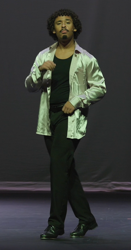 A man with curly hair standing on a stage, wearing a satin-like silver shirt, black undershirt, black pants, and black shoes.