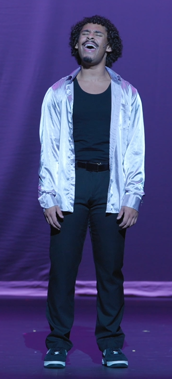A man singing passionately on stage with eyes closed, wearing a pink satin shirt, white undershirt, black pants, and sneakers, against a purple backdrop.