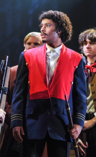 A young man in a theatrical costume in 'Les Miserables', surrounded by other performers, on stage.