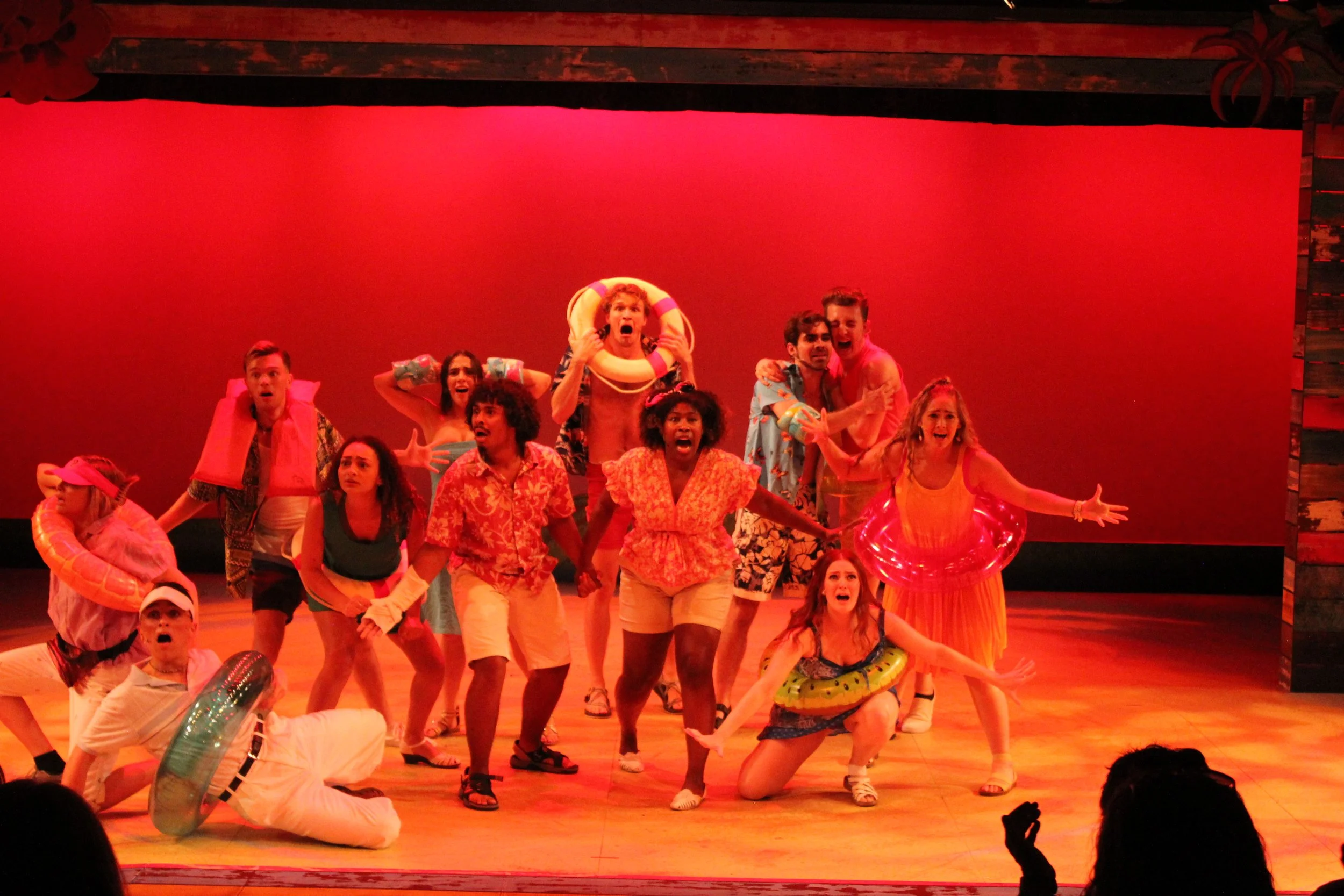 The cast of a musical or play on stage dressed in summer beachwear, including swimsuits, shorts, and tropical shirts, with some wearing swimming rings. The background is a solid red, and the performers express surprise, excitement, or joy.