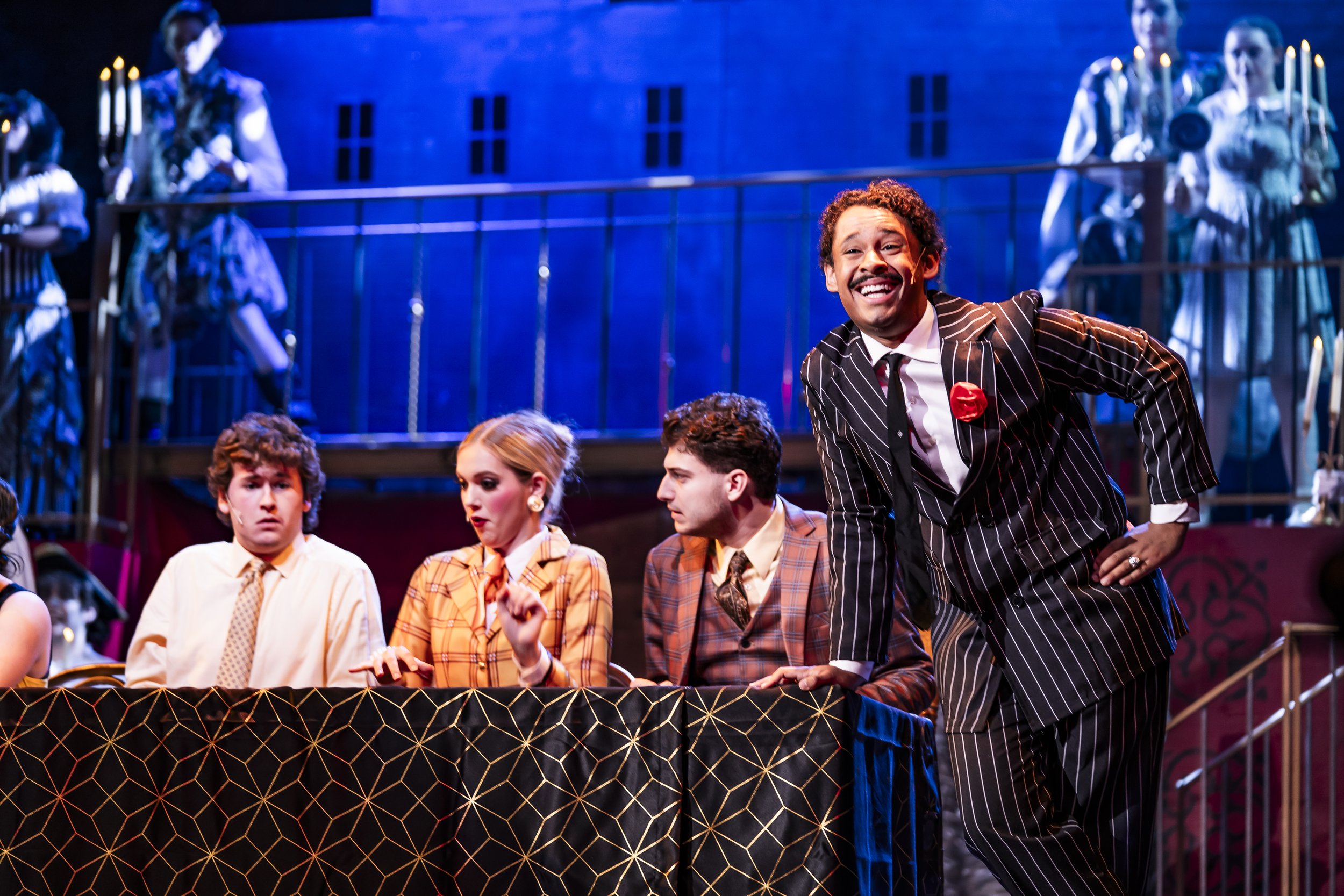 Actors performing 'The Addams Family' musical with some seated at a table and one standing, dressed in vintage-style clothing, in a colorful, theatrical setting with costumes and set pieces in the background.