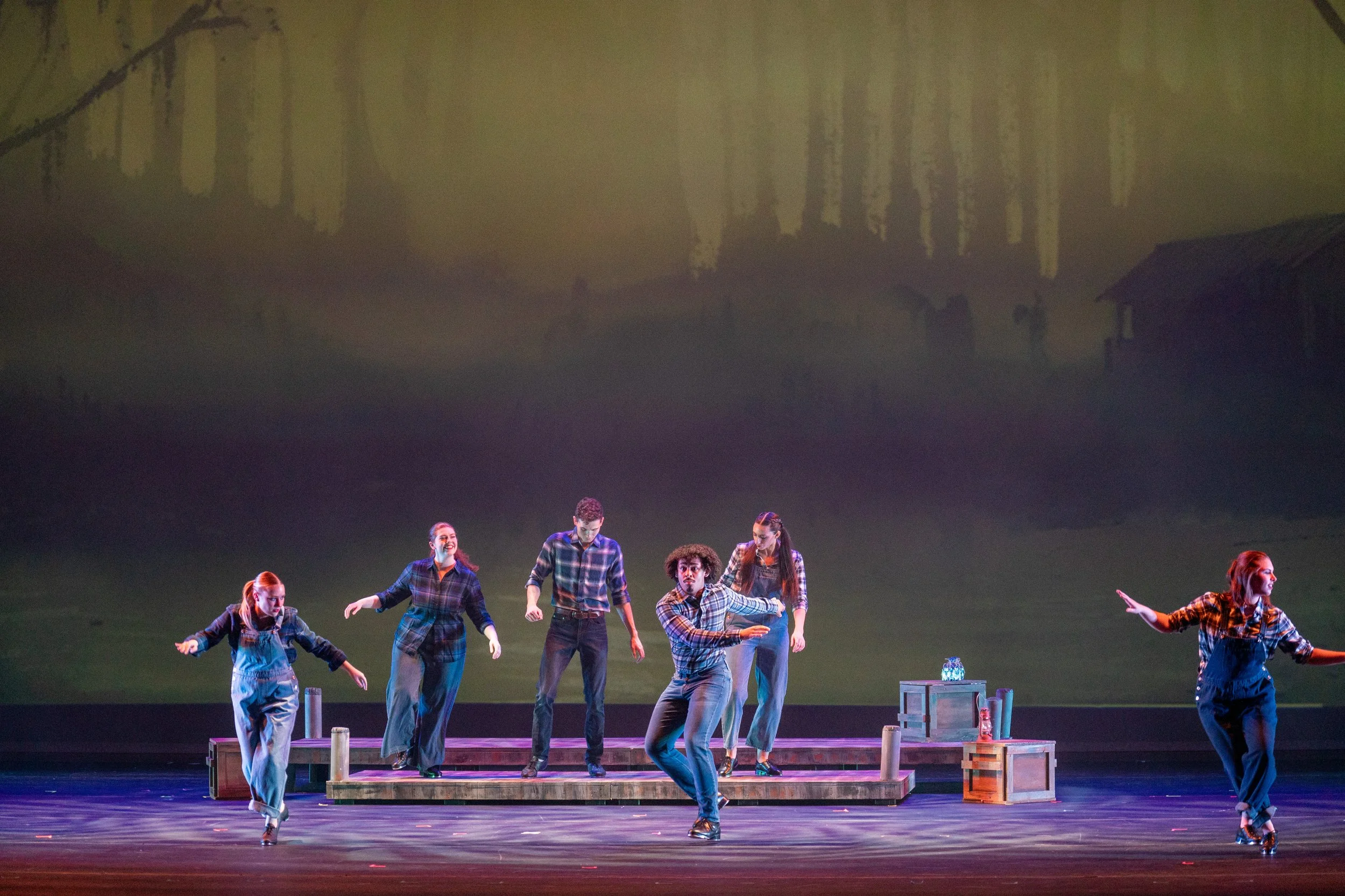 A theatrical stage performance with six dancers dressed in plaid shirts and jeans, performing a tap routine. The stage has a set with crates and bottles, and a backdrop showing a countryside scene with trees and a house.