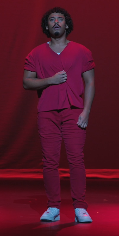 A man person with curly hair in a red shirt and pants standing on a stage with a red background.