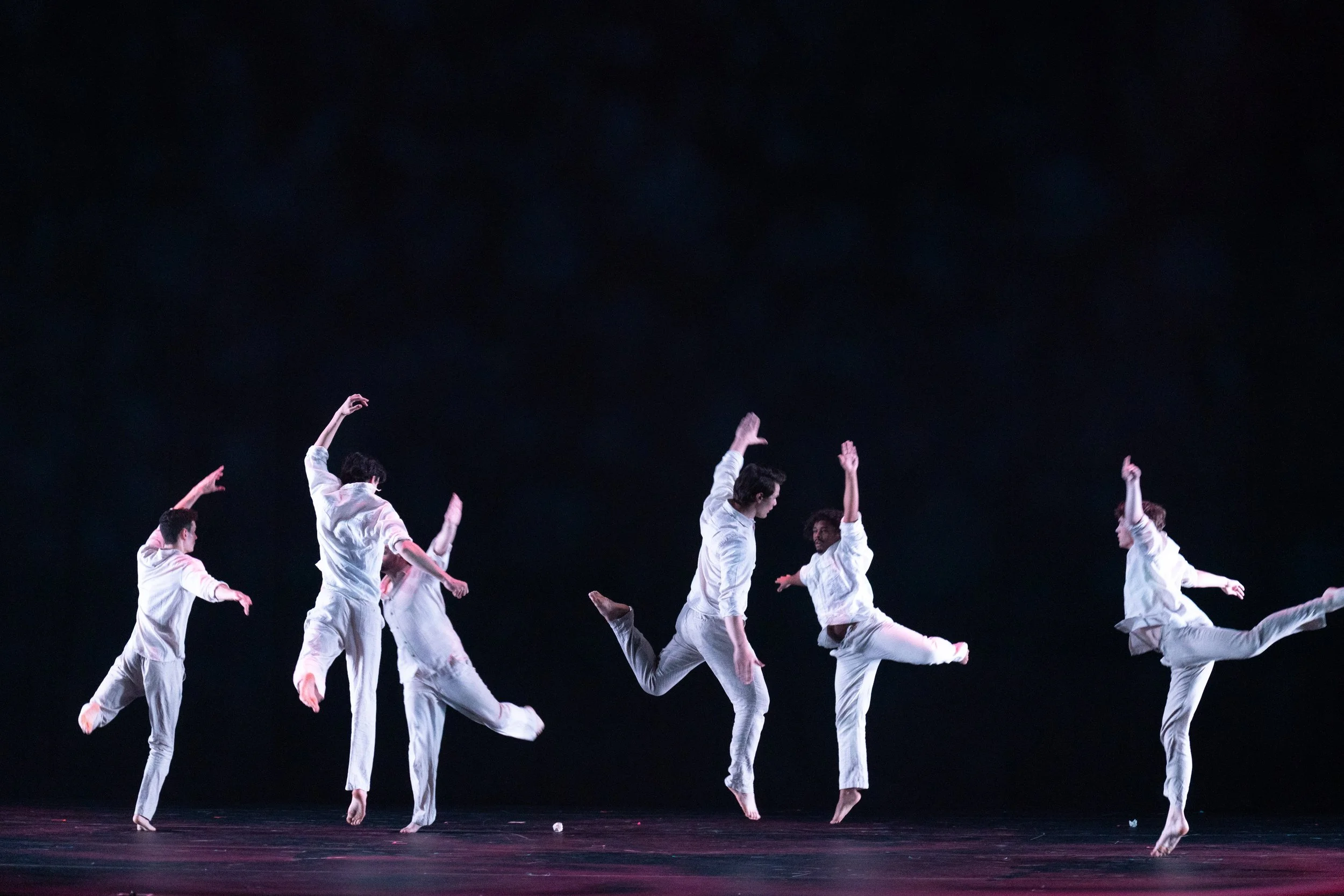 Six dancers in white outfits performing a modern dance routine on stage against a black background.