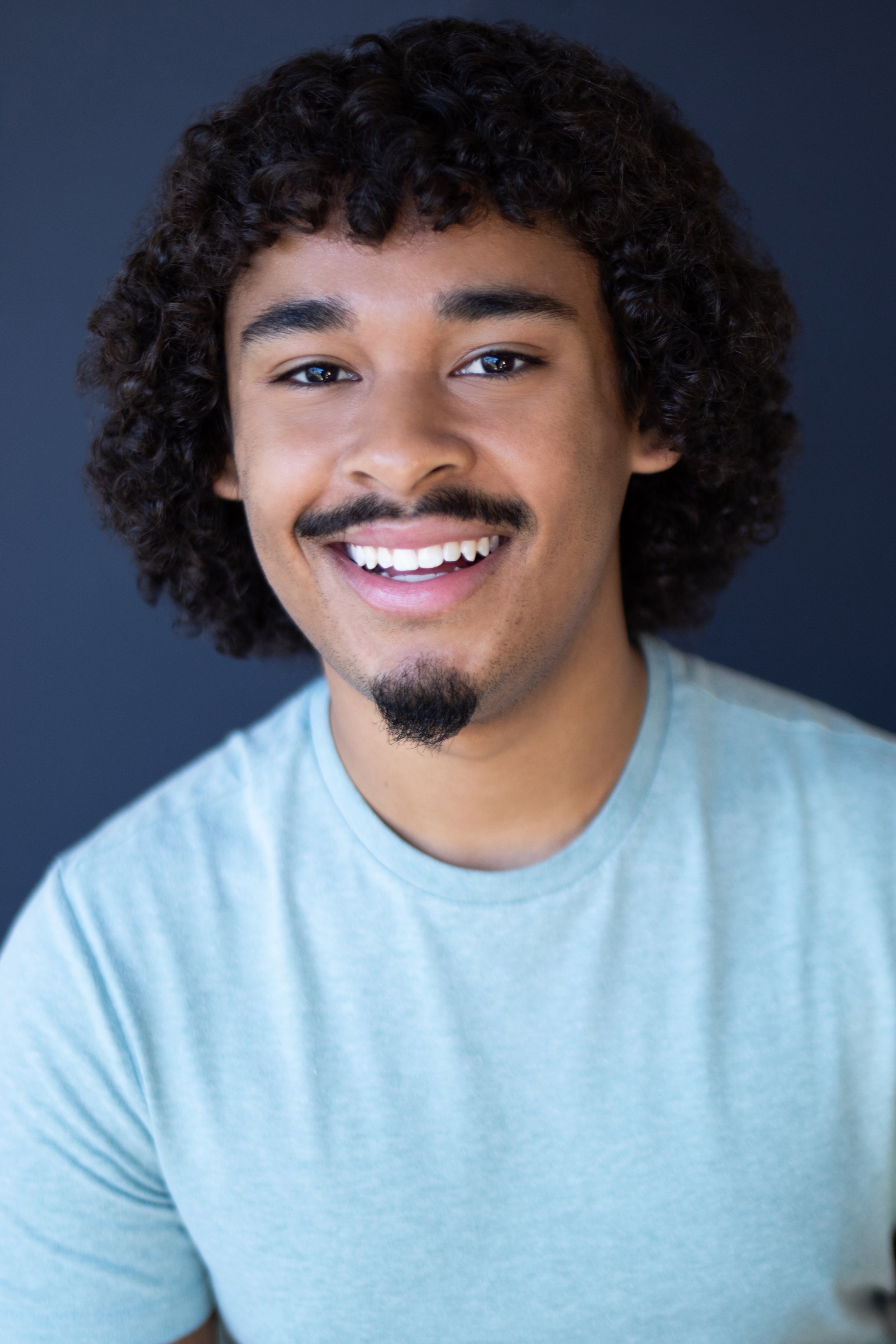 Portrait of a handsome young man with curly hair and a goatee, wearing a light green shirt, smiling against a dark background.