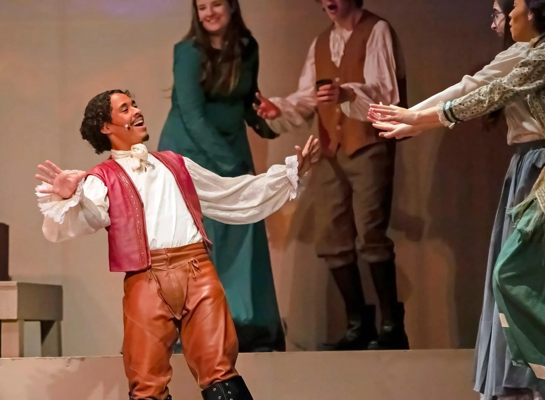 Stage scene with five actors. One male actor with curly hair, as Shakespeare in 'Something Rotten!' musical, dressed in a historical costume with leather pants, a white shirt, and a red vest, smiling and gesturing with his hands. 