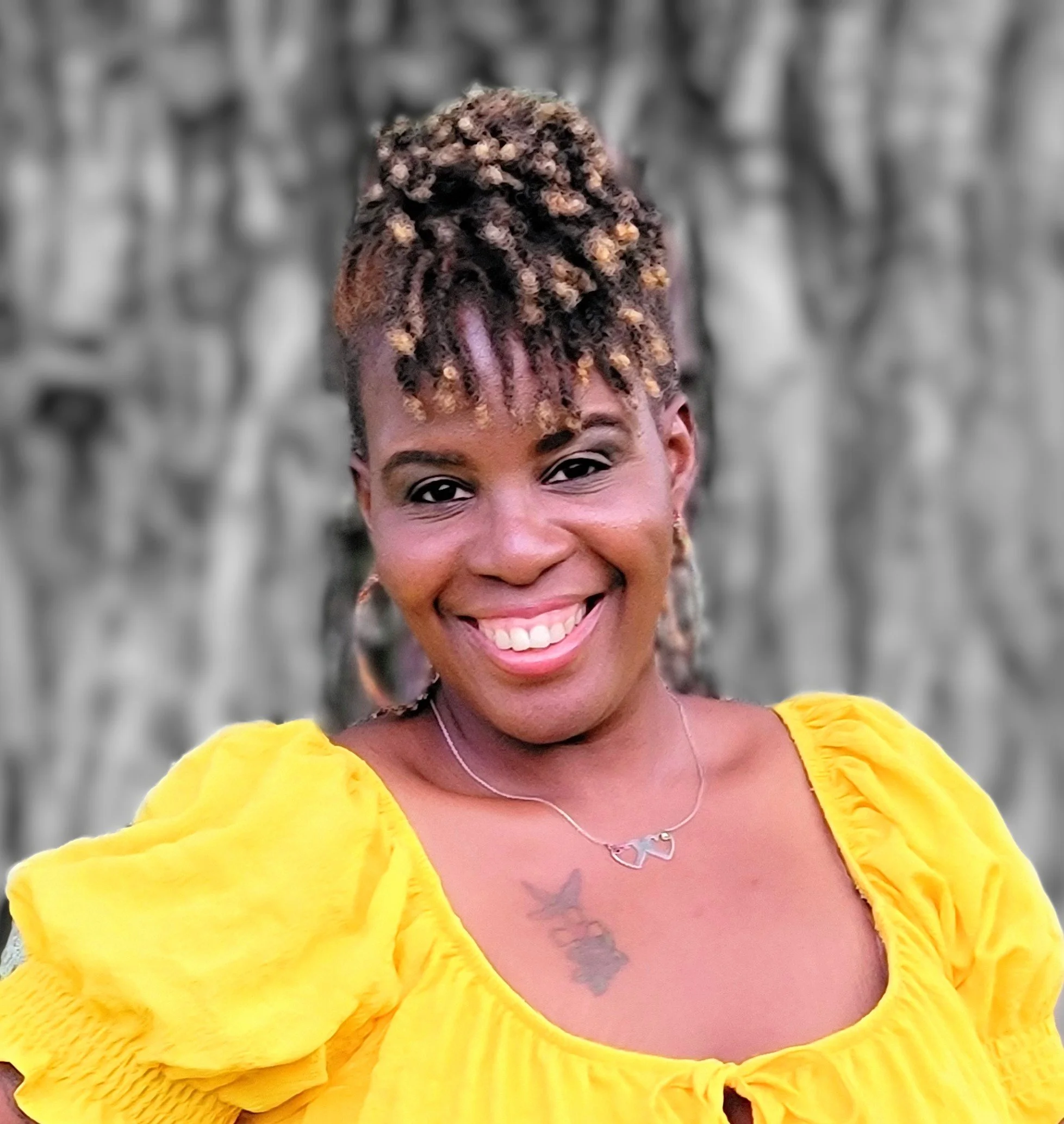A smiling woman with natural hair styled in short, twisted locks with blonde tips, wearing a yellow blouse with puffed sleeves, a butterfly necklace, and earrings, standing outdoors in front of a blurred gray stone wall.
