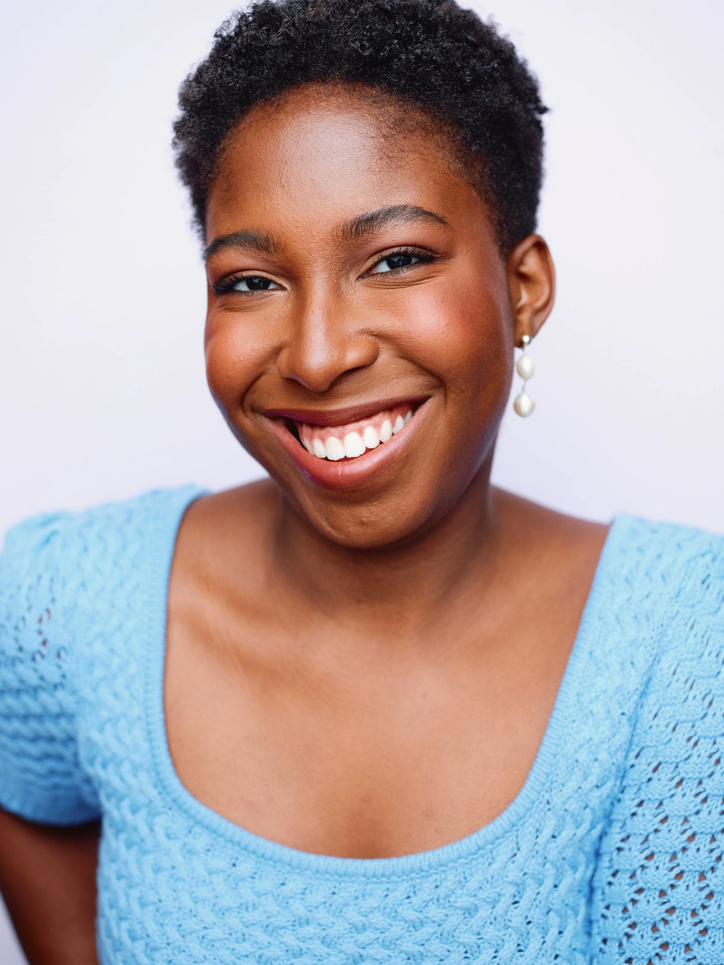 A woman smiling with short, curly hair, wearing pearl earrings and a light blue knitted top.