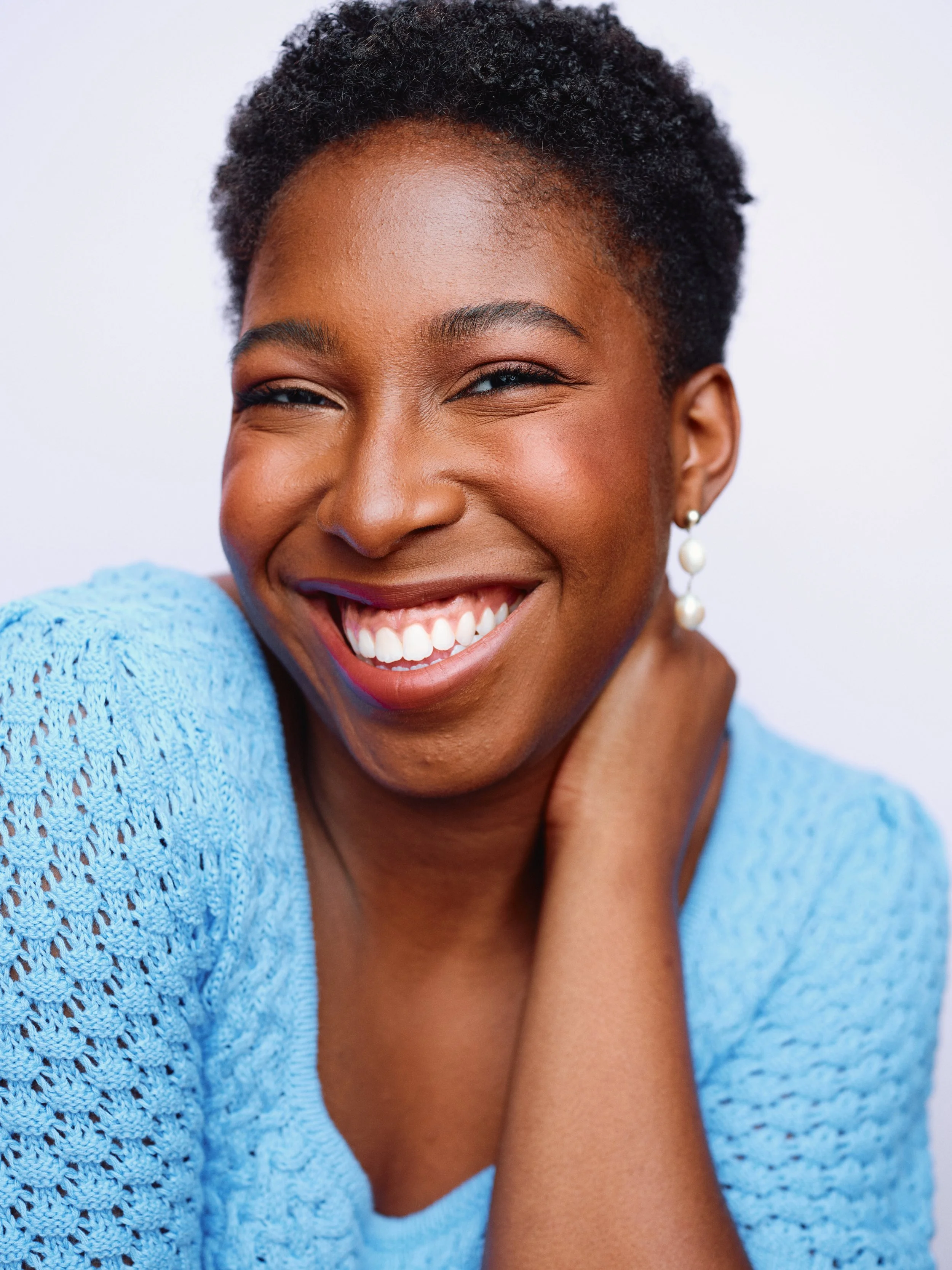 Close-up of a woman with short curly hair smiling brightly, wearing a light blue knitted top and pearl earrings.