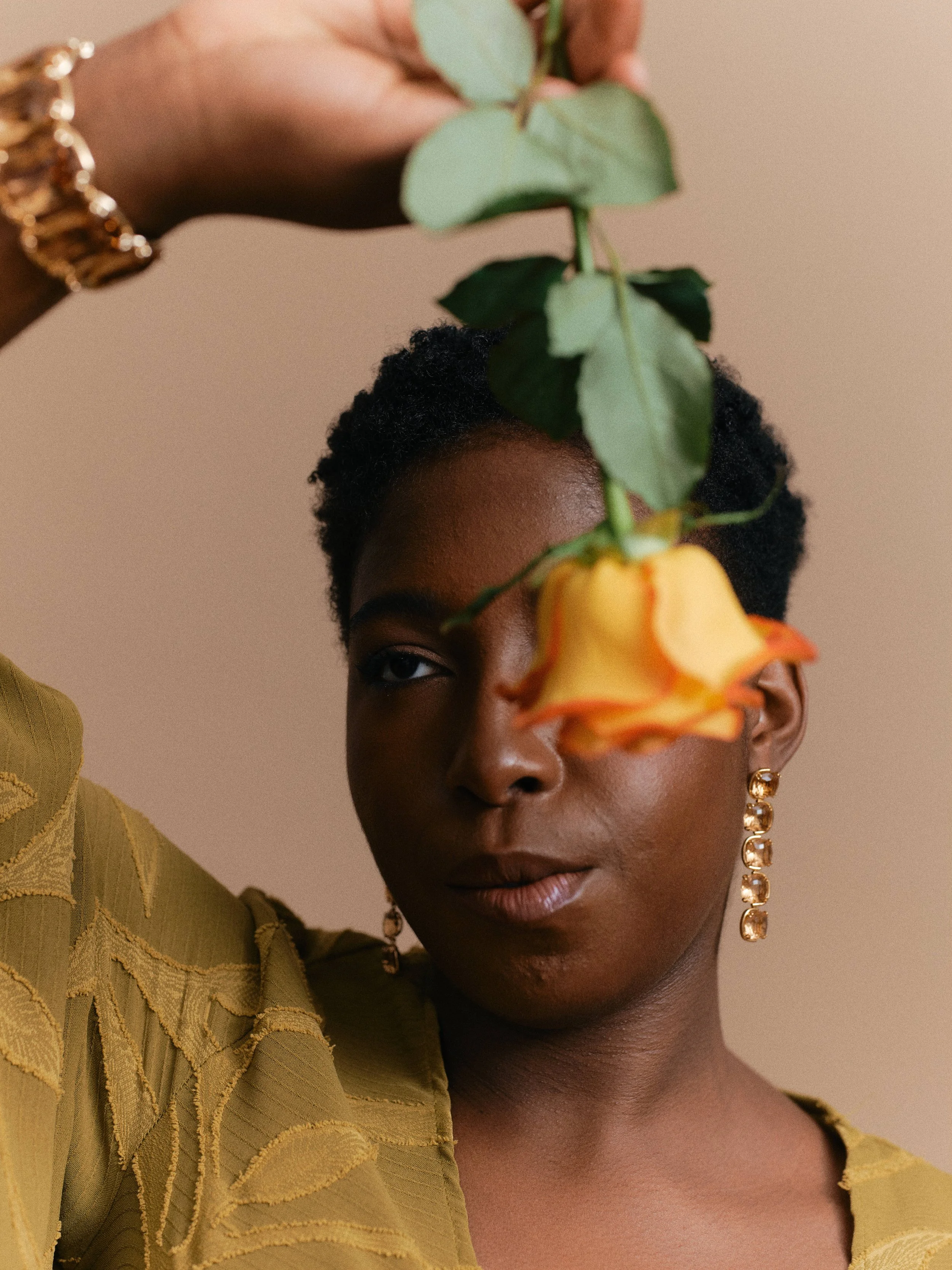 A woman with dark skin and short curly hair holding a yellow and orange rose in front of her face. She is wearing gold earrings and a gold bracelet. Her expression is calm, and she is wearing a textured yellow-green top.