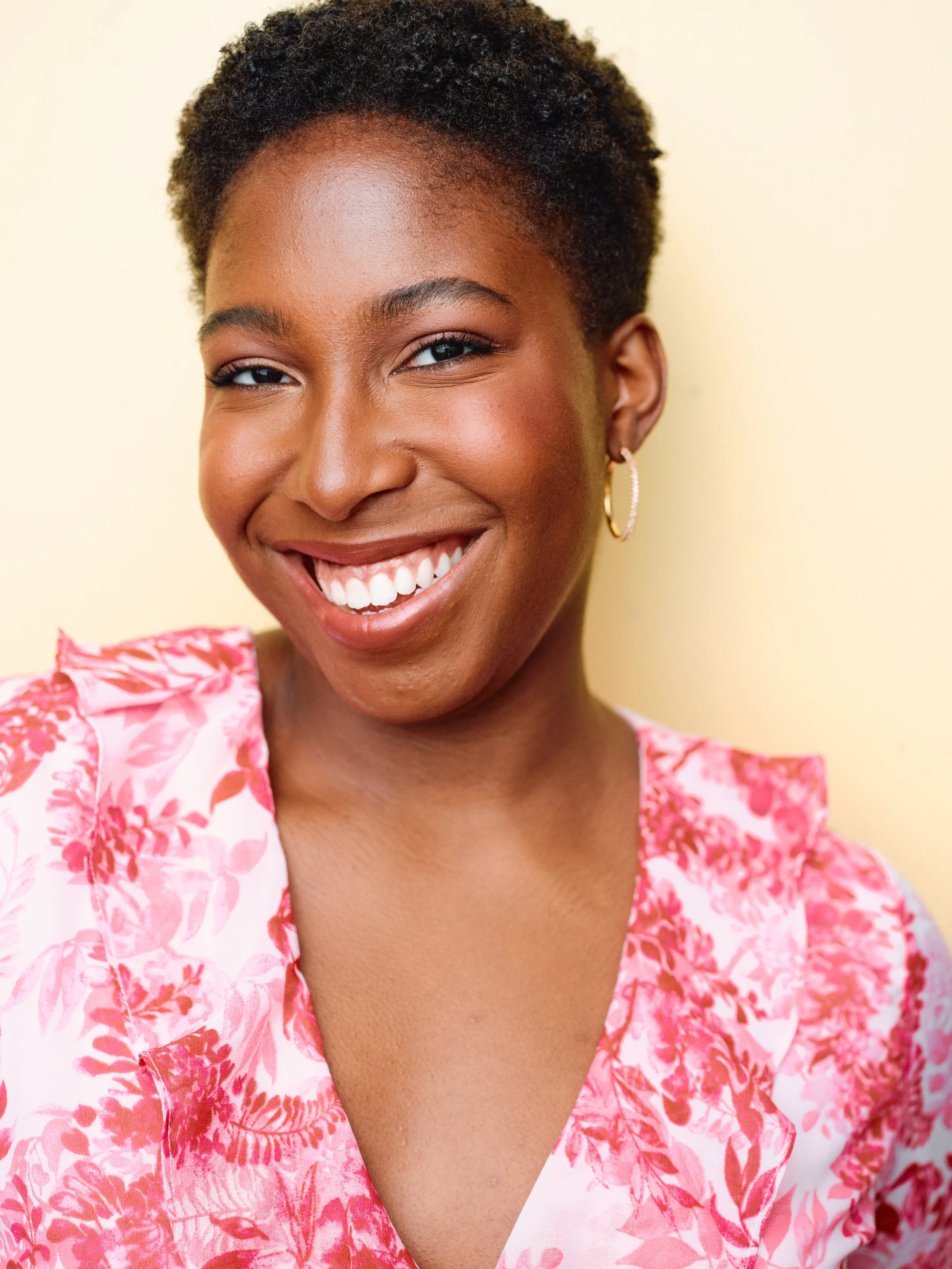 A woman with short, curly hair smiling, wearing a pink and white floral top and hoop earrings, against a light yellow background.