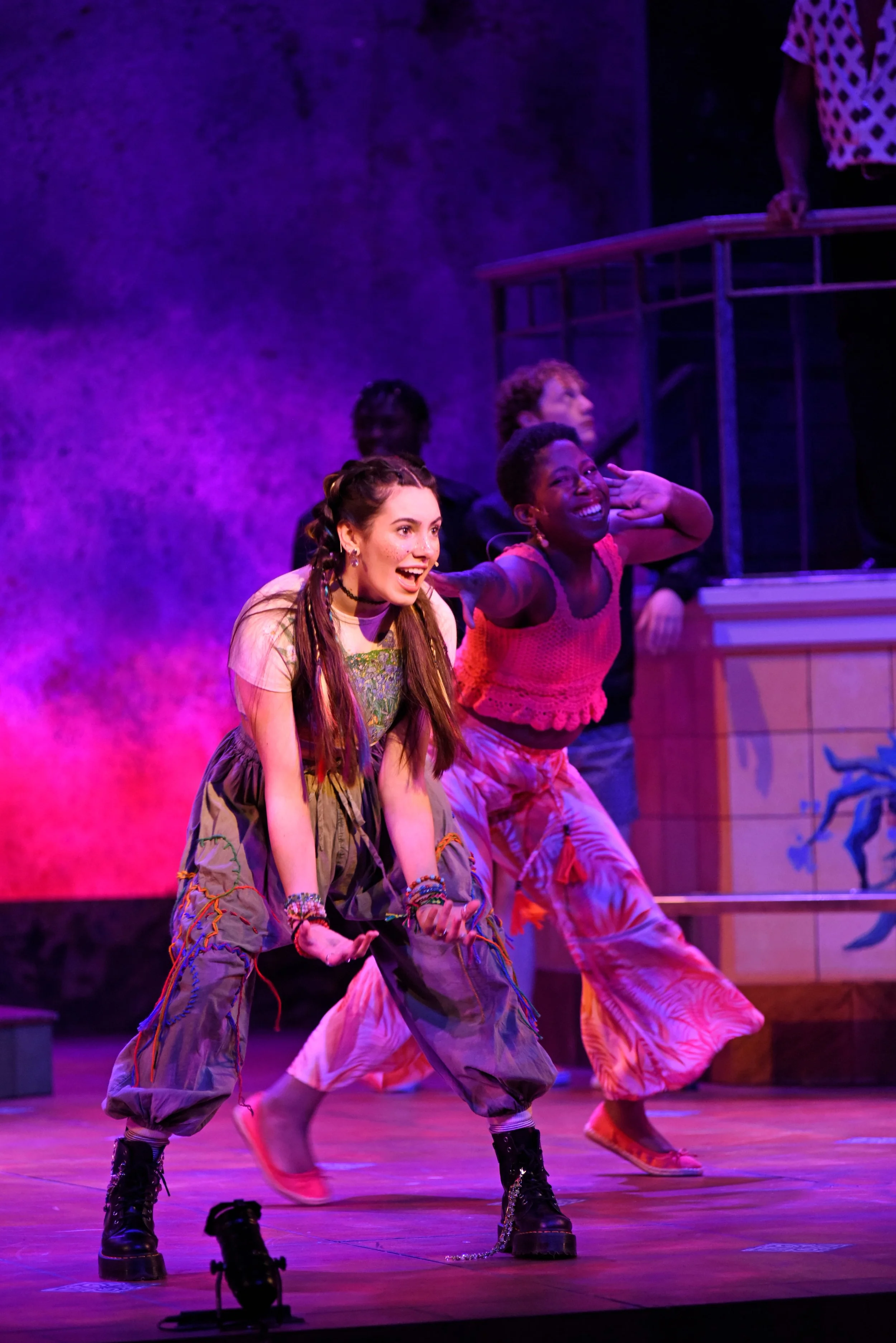 Two young women perform a lively dance on stage, wearing colorful casual outfits and smiling in front of a vibrant purple and pink background.