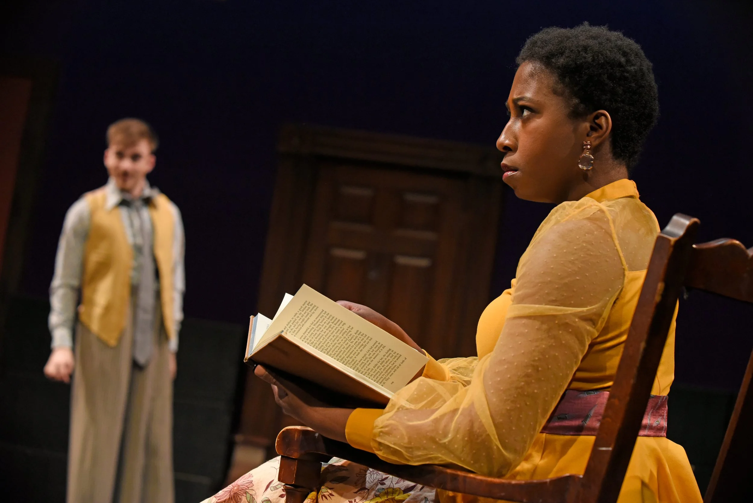 A woman dressed in a yellow dress and sheer yellow sleeves is sitting in a wooden chair, reading a book with a serious expression. In the background, a young man in vintage clothing appears surprised or upset.